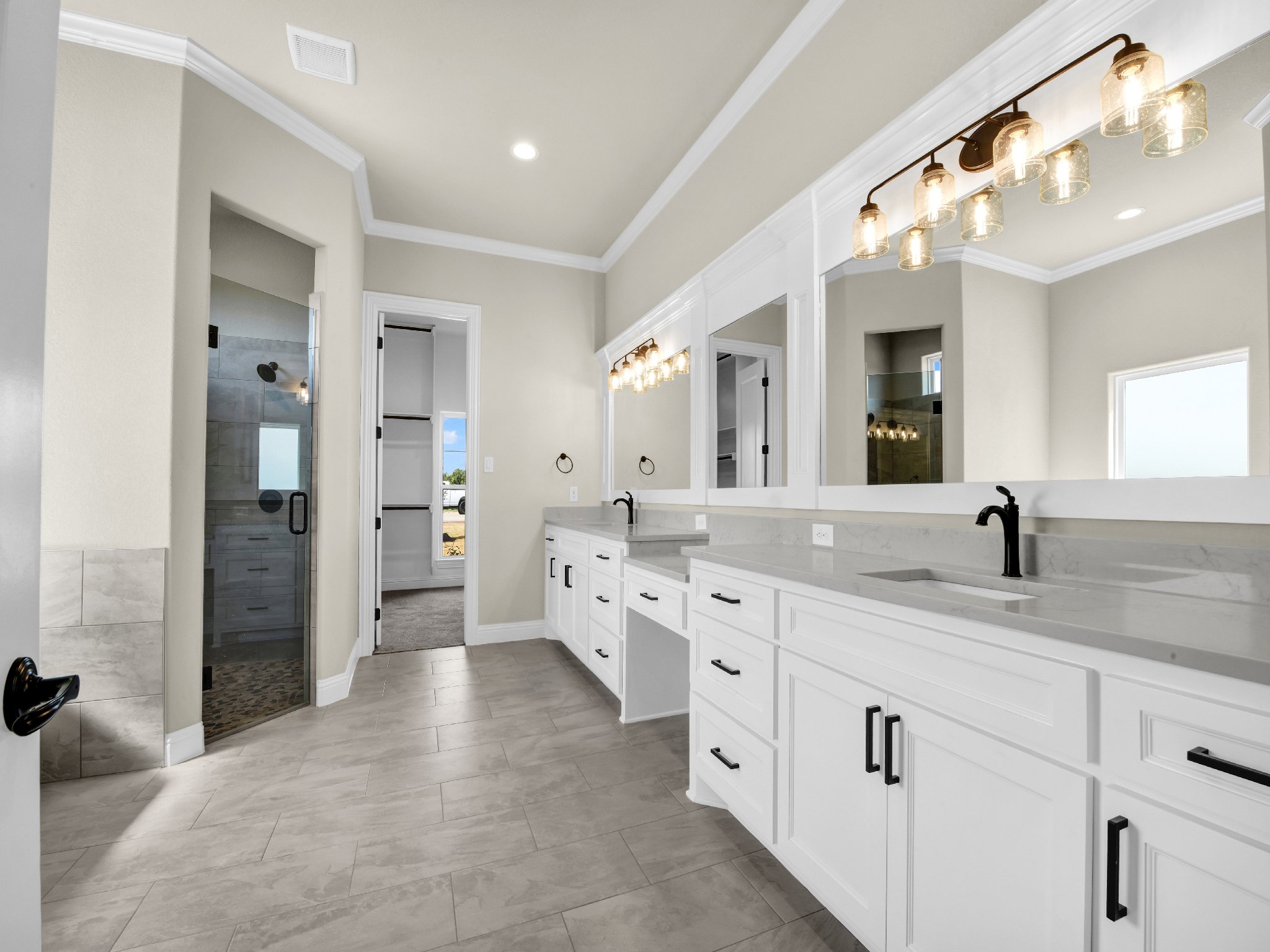 Modern bathroom with white cabinets, black handles, gray countertops, and a large mirror, with a walk-in shower and windows providing natural light.