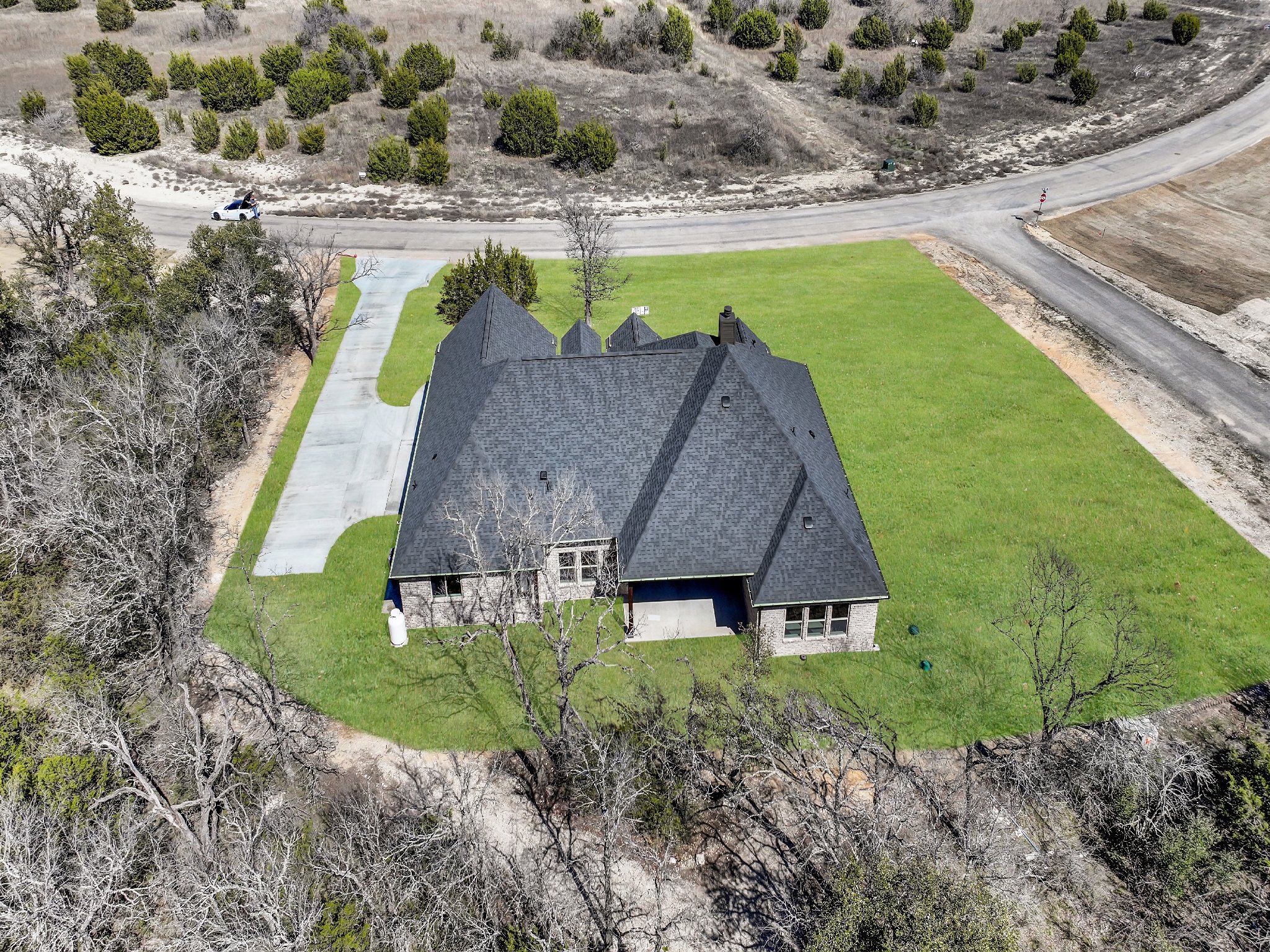 Aerial view of a house with gray roof and brick walls, surrounded by a well-maintained lawn and leafless trees, with gravel driveways and dirt roads nearby.