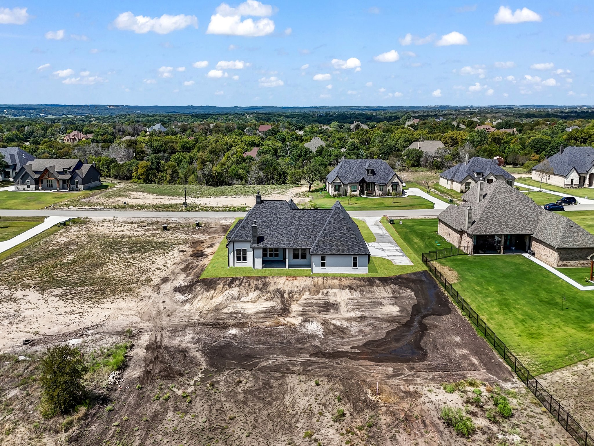 Aerial view of a residential neighborhood with several houses, some with finished exteriors and some under construction, with a partially developed lot and green lawns on a sunny day with scattered clouds.