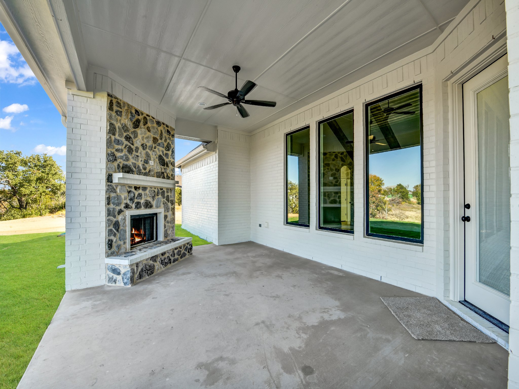 Covered outdoor patio with a stone fireplace, large windows, and a ceiling fan, overlooking a grassy yard with trees.