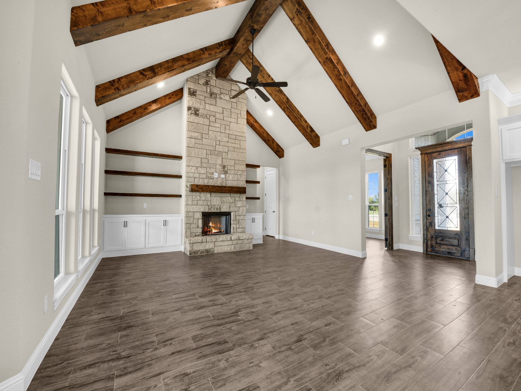 Empty living room with wooden floors, beige walls, and a stone fireplace with built-in shelves on each side. The ceiling has exposed wooden beams and a ceiling fan. There is a rustic front door with glass panels and a small window next to it.