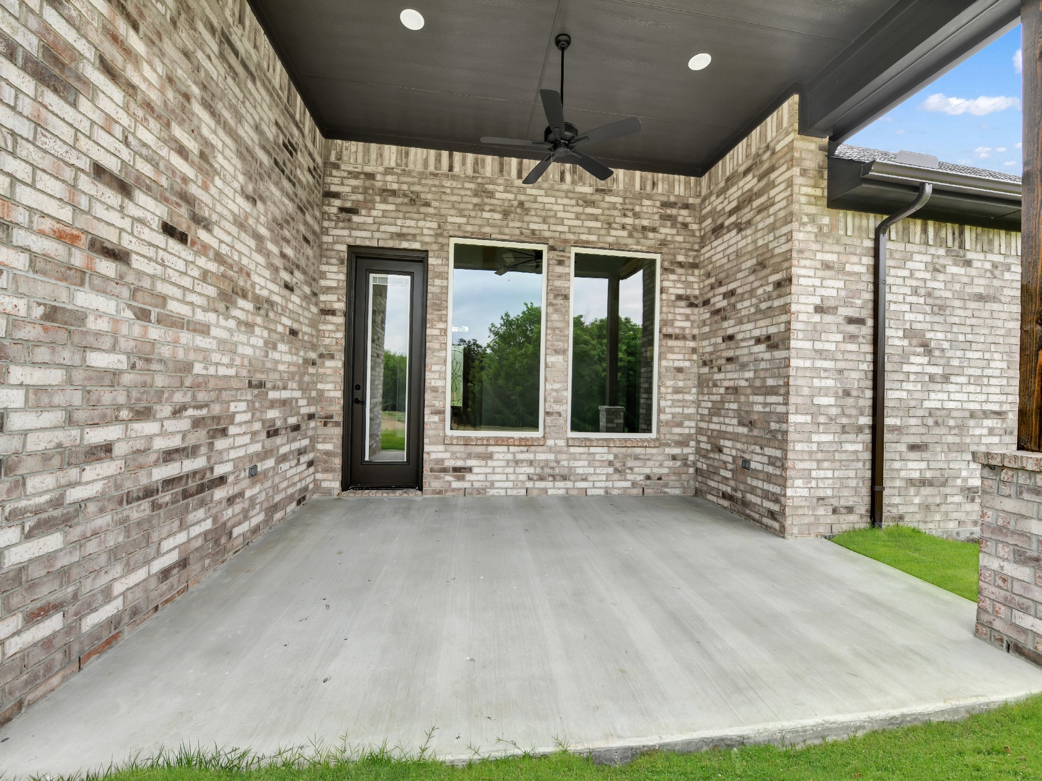 Empty outdoor covered patio with brick walls, concrete floor, black door and windows, ceiling fan, and green grass outside.