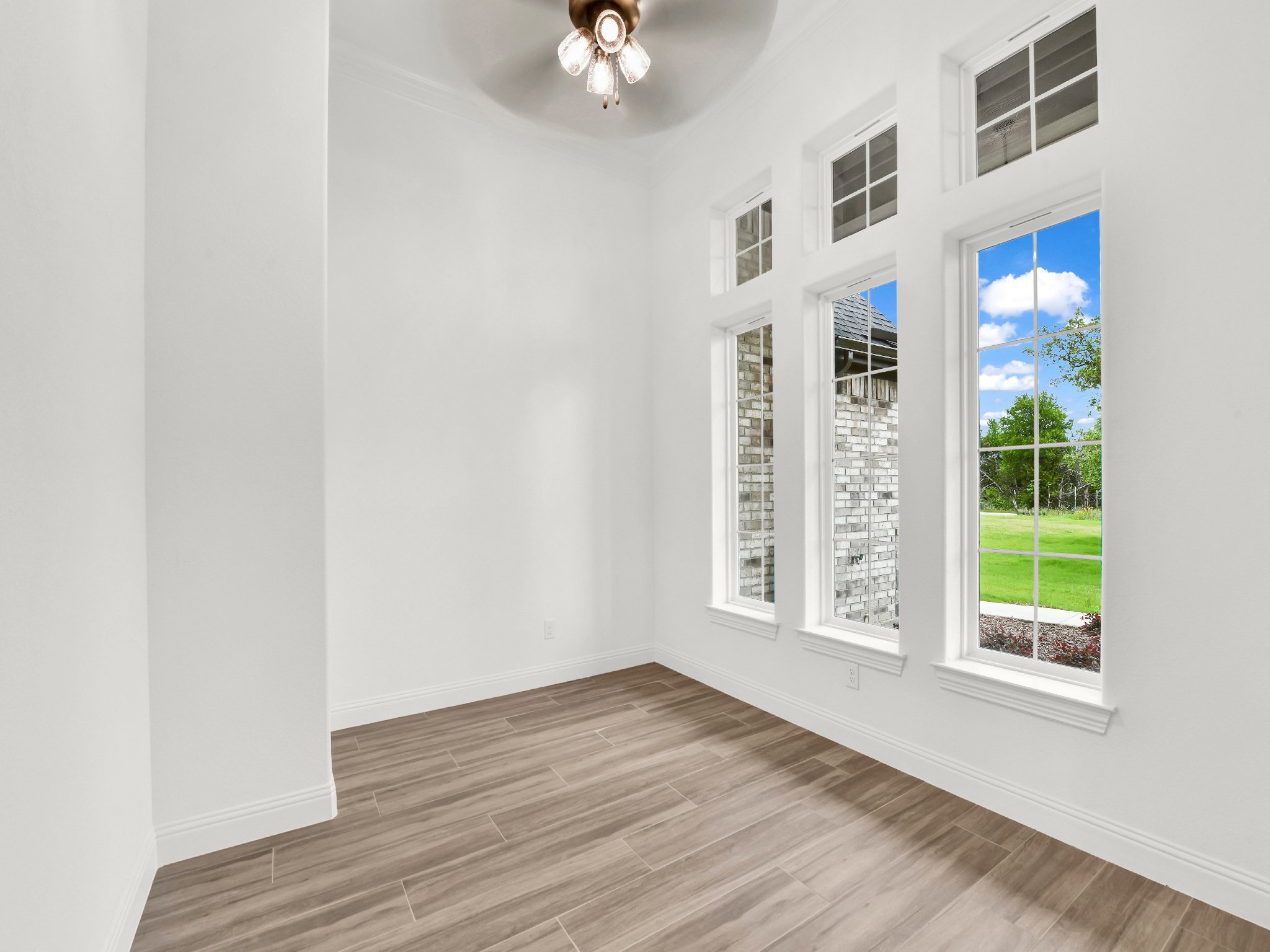 Empty room with large windows, white walls, wood flooring, and a ceiling fan with lights, overlooking a green lawn and blue sky outside.