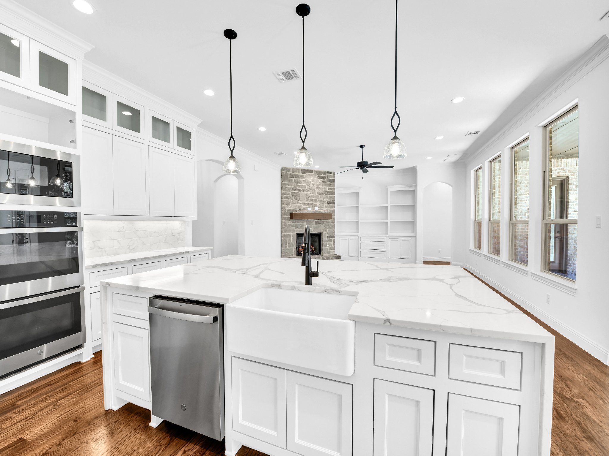 Modern white kitchen with marble countertops, built-in appliances, black fixtures, hardwood floors, large windows, and a fireplace in the living area.