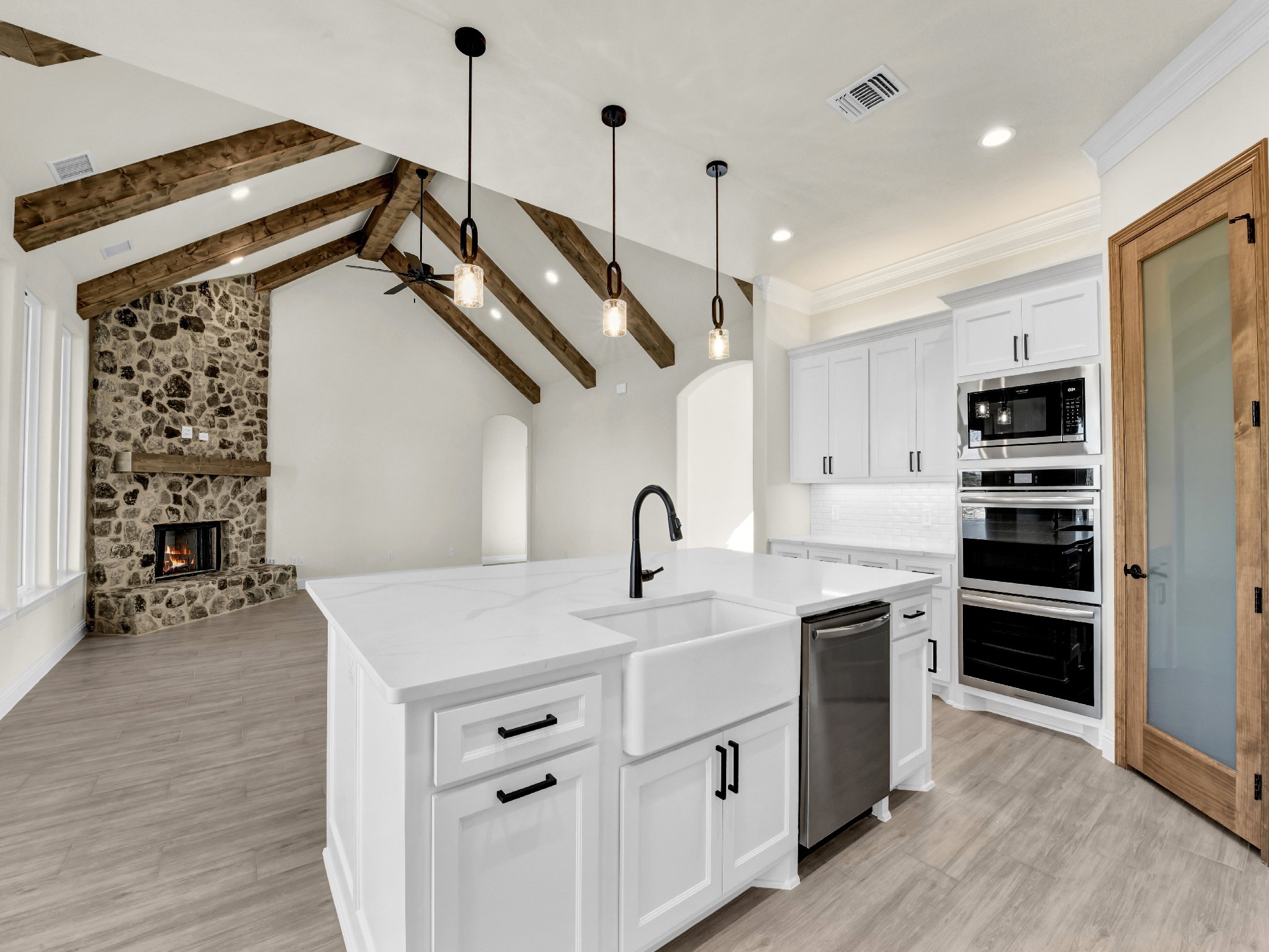 Modern kitchen with white cabinets, black handles, stainless steel appliances, a white island with a black faucet, wooden beams on the ceiling, and a stone fireplace in an open living area.