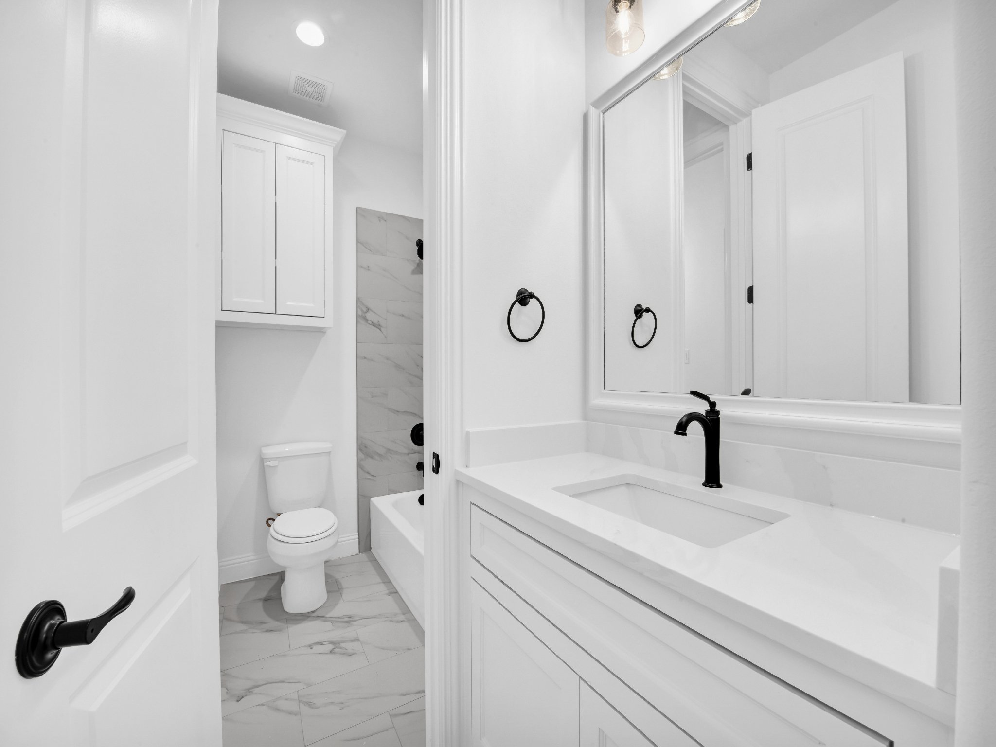 Modern white bathroom with black fixtures, including sink faucet and towel rings, large mirror, and a separate toilet and shower area with marble walls.