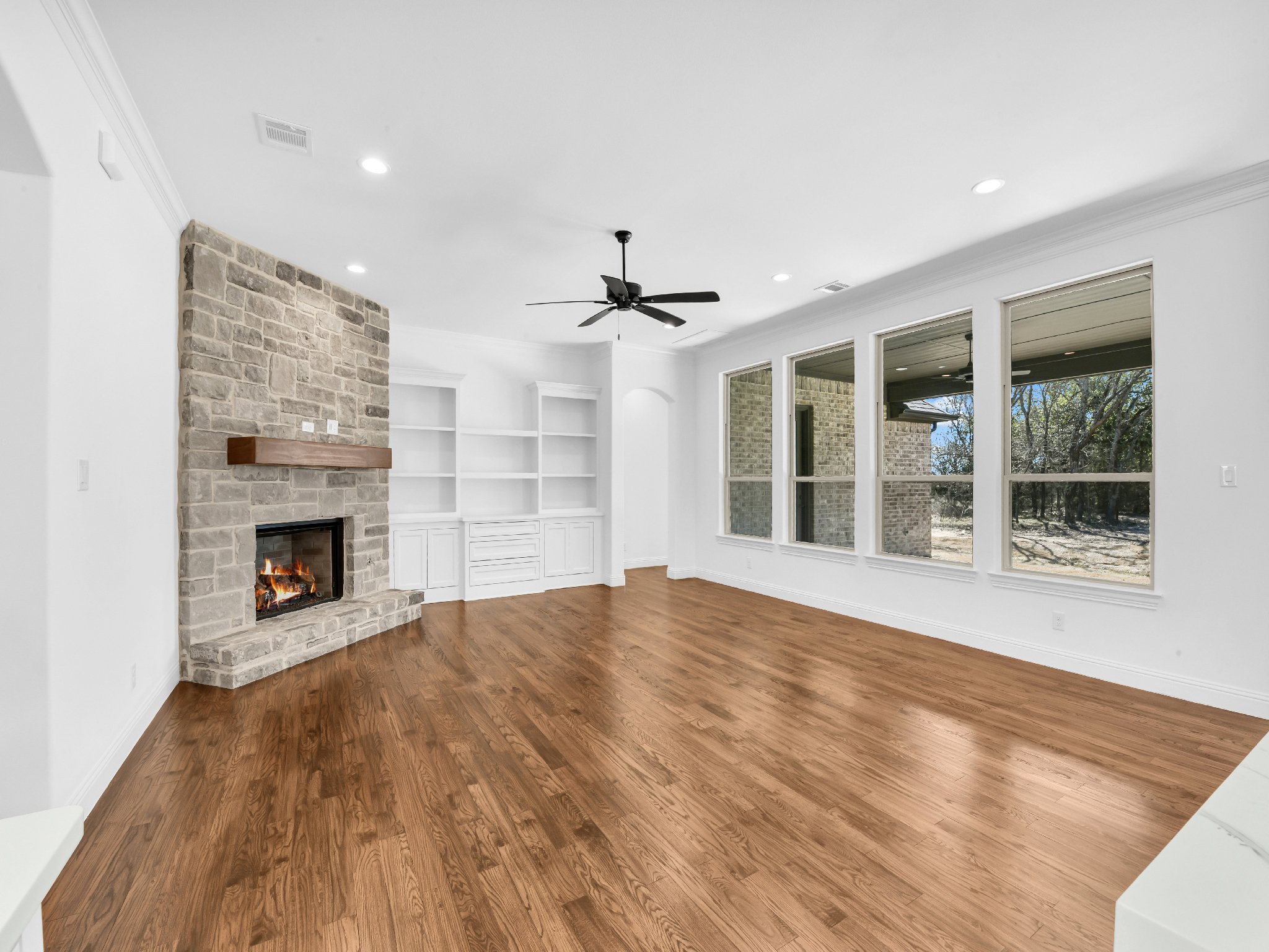 Empty living room with vinyl plank flooring, a stone fireplace with a wooden mantel, white built-in shelves and cabinets, large windows with a view of trees outside, a ceiling fan, and recessed lighting.