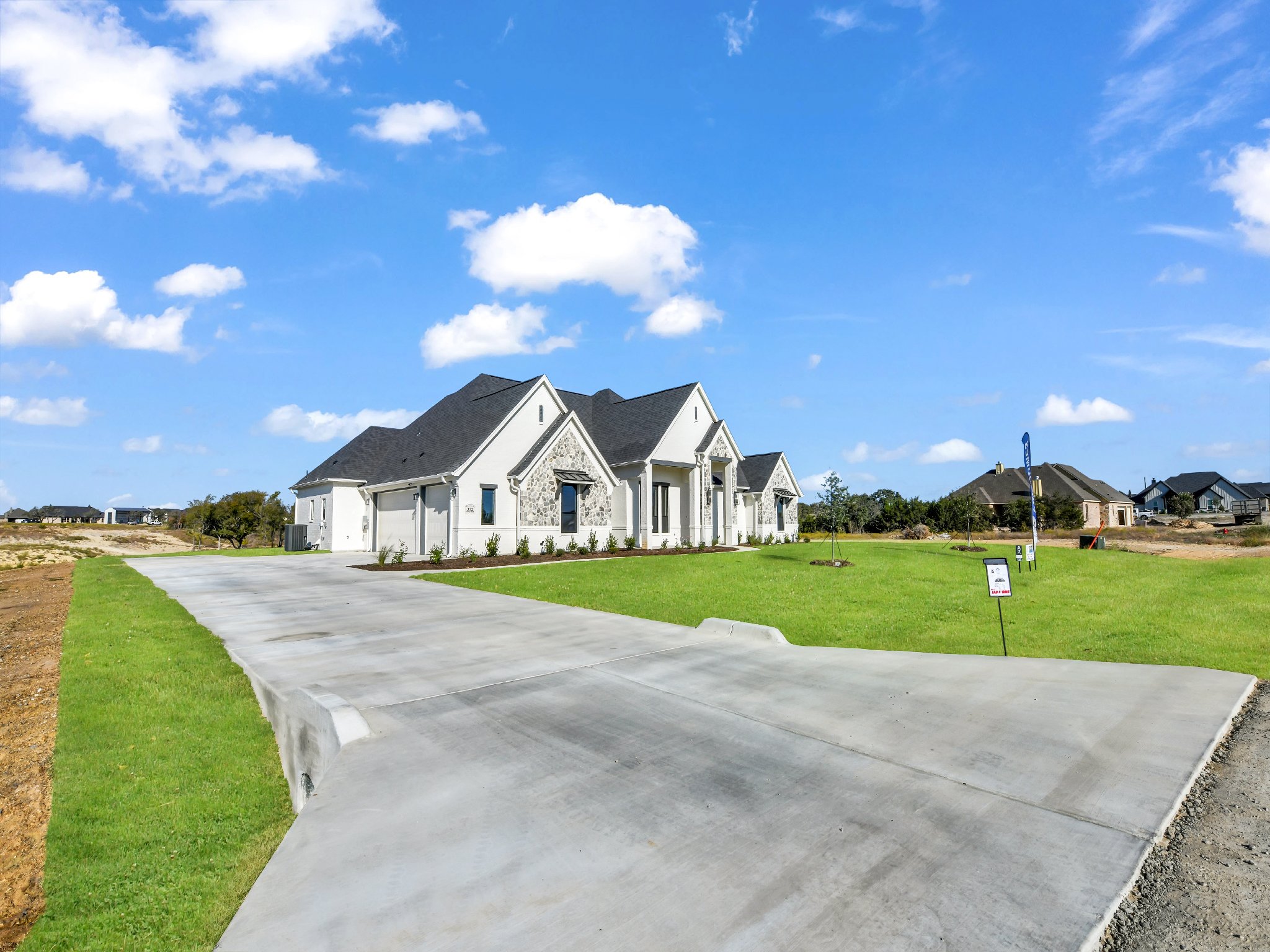 Newly built white house with gray roof and stone accents, green lawn, and a curved concrete driveway, under a blue sky with scattered clouds.