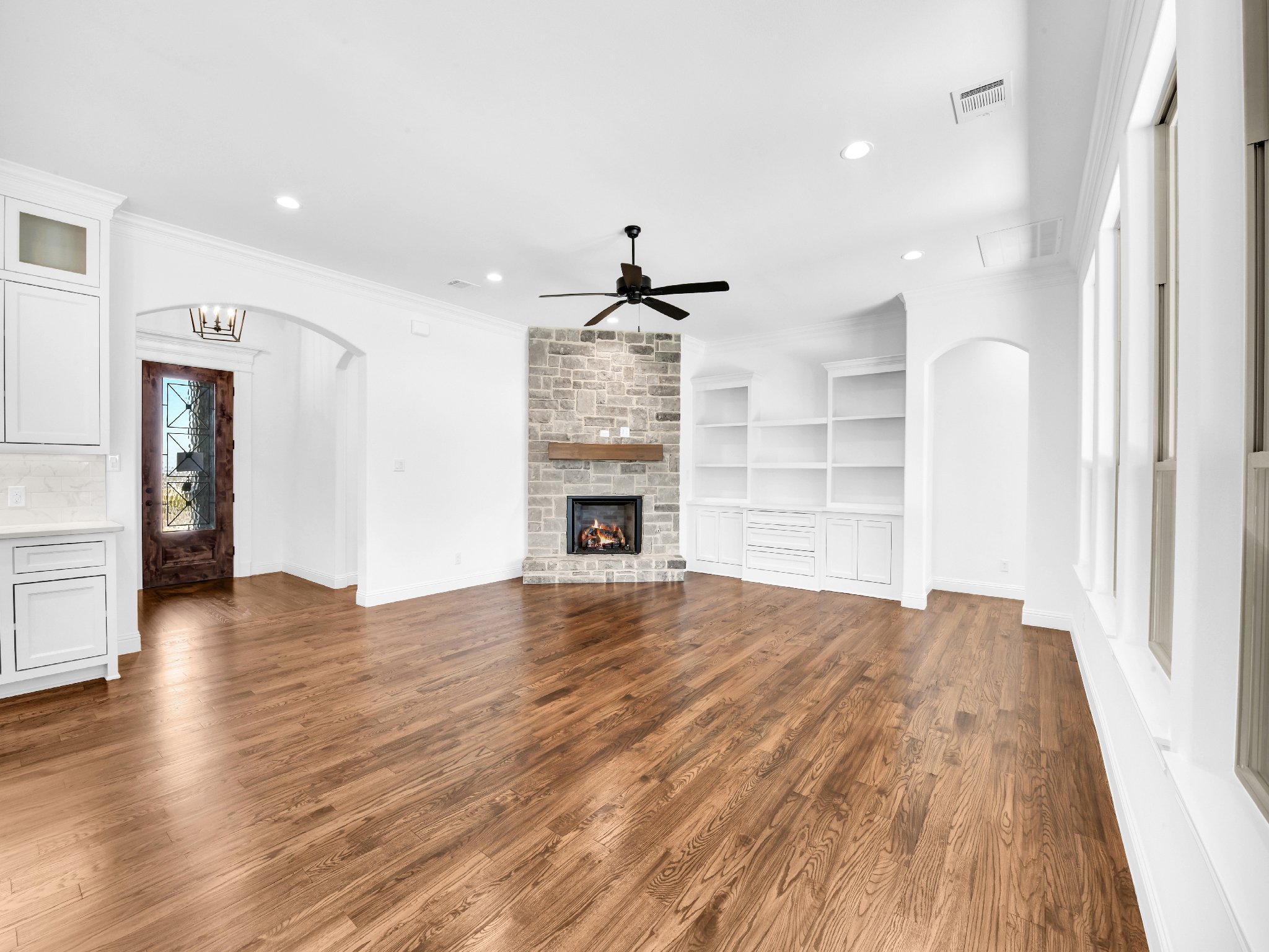 Living room with white walls, hardwood floors, a stone fireplace, white built-in shelves, large windows, and a ceiling fan.