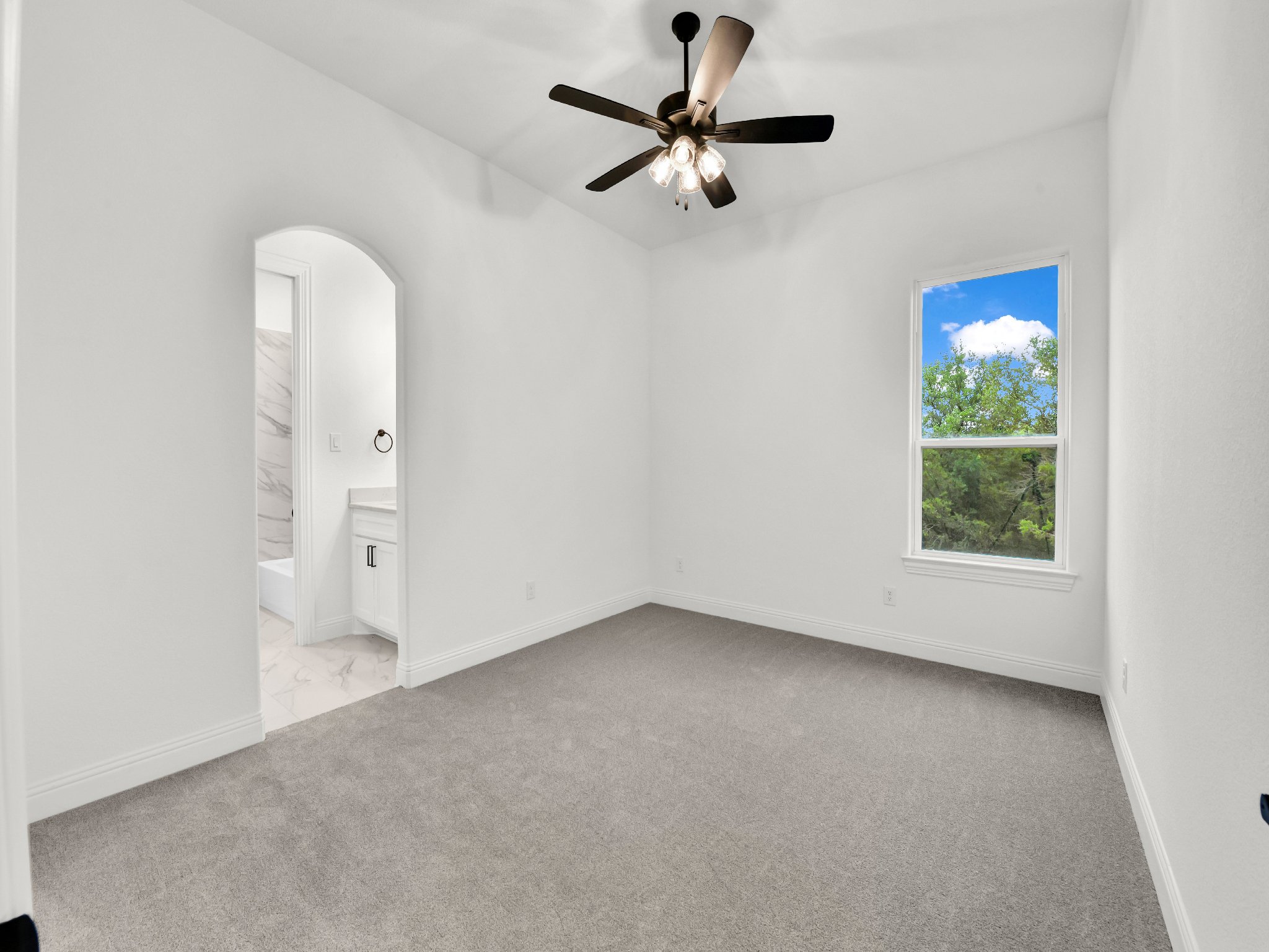 Empty bedroom with white walls, a window showing greenery outside, a ceiling fan with lights, and an attached bathroom with marble walls