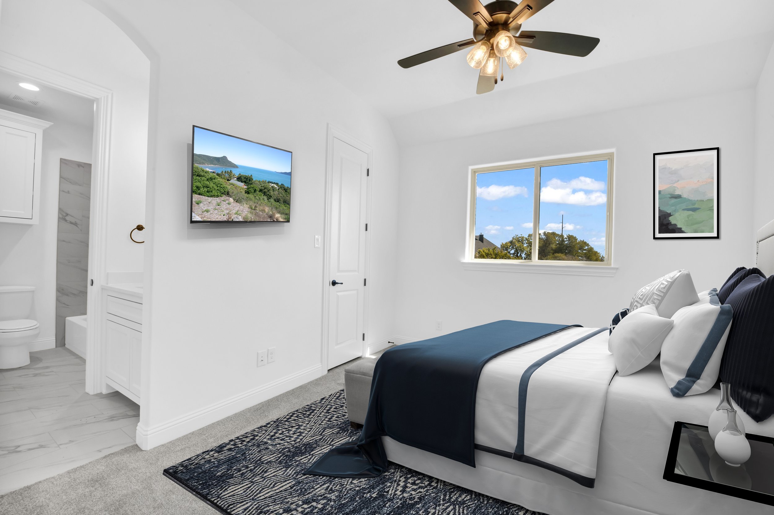 Bright bedroom with white walls, black and white bedding, window with view of trees and blue sky, ceiling fan, wall art, flat-screen TV, and an adjacent bathroom.