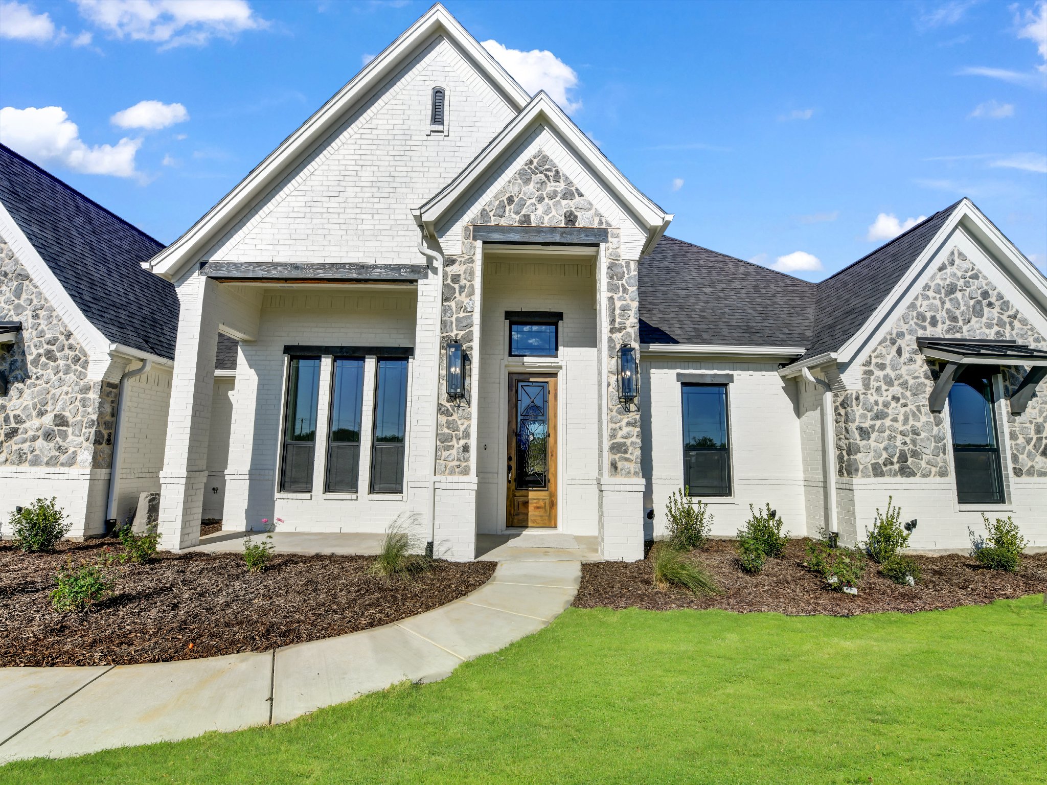 A modern two-story house with white brick and gray stone exterior, black-framed windows, and a front door, surrounded by a green lawn and a curved stone walkway.