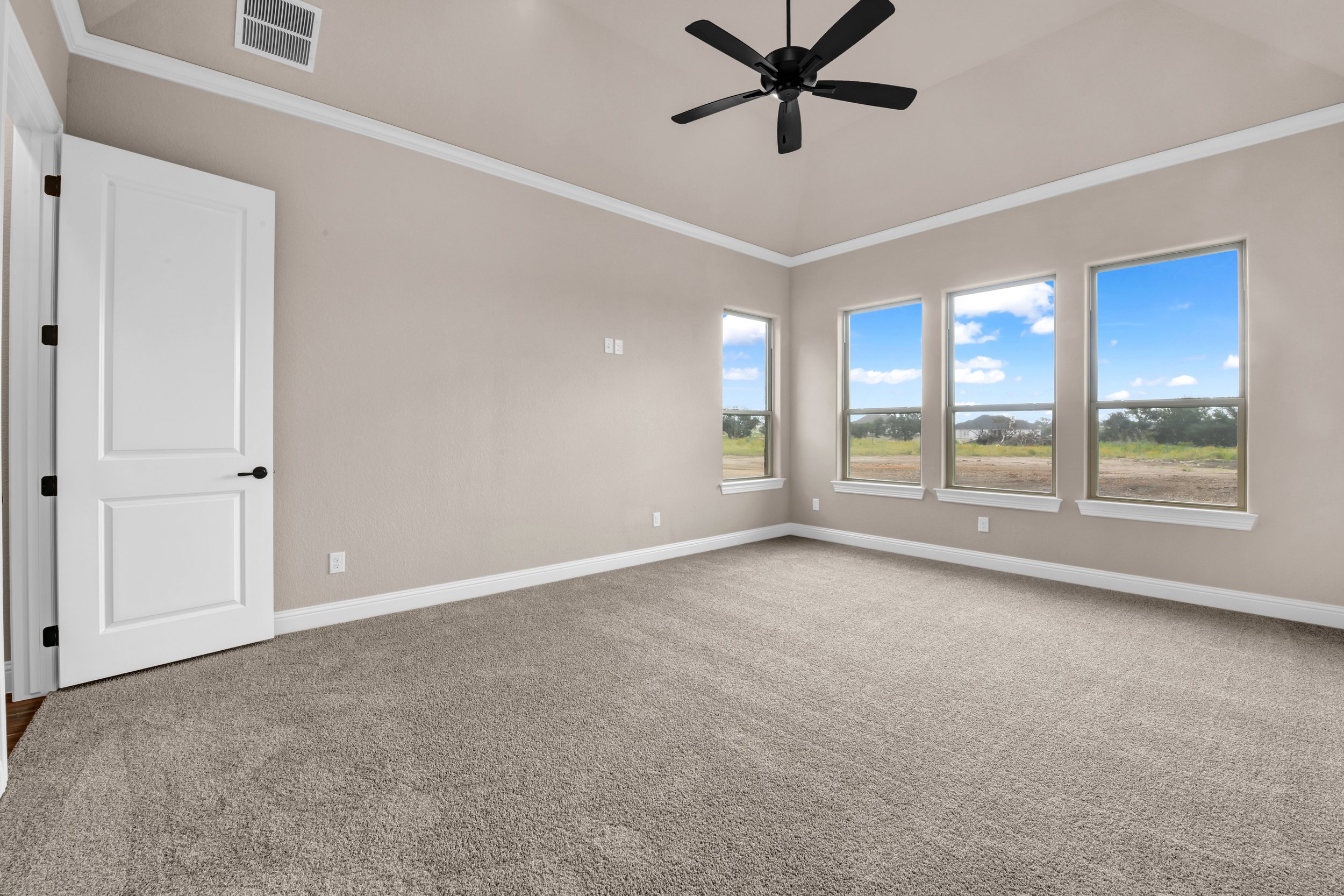Empty room with beige walls, carpeted floor, four large windows showing a blue sky and outdoor landscape, a ceiling fan, and white trim.