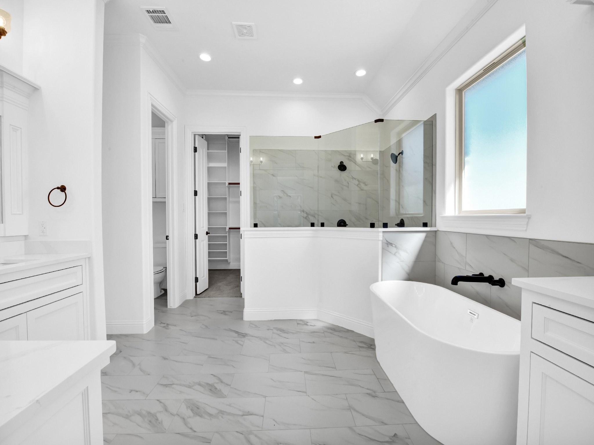 Bright white bathroom with marble floors and wall accents, freestanding bathtub, large window, and a glass-enclosed shower.