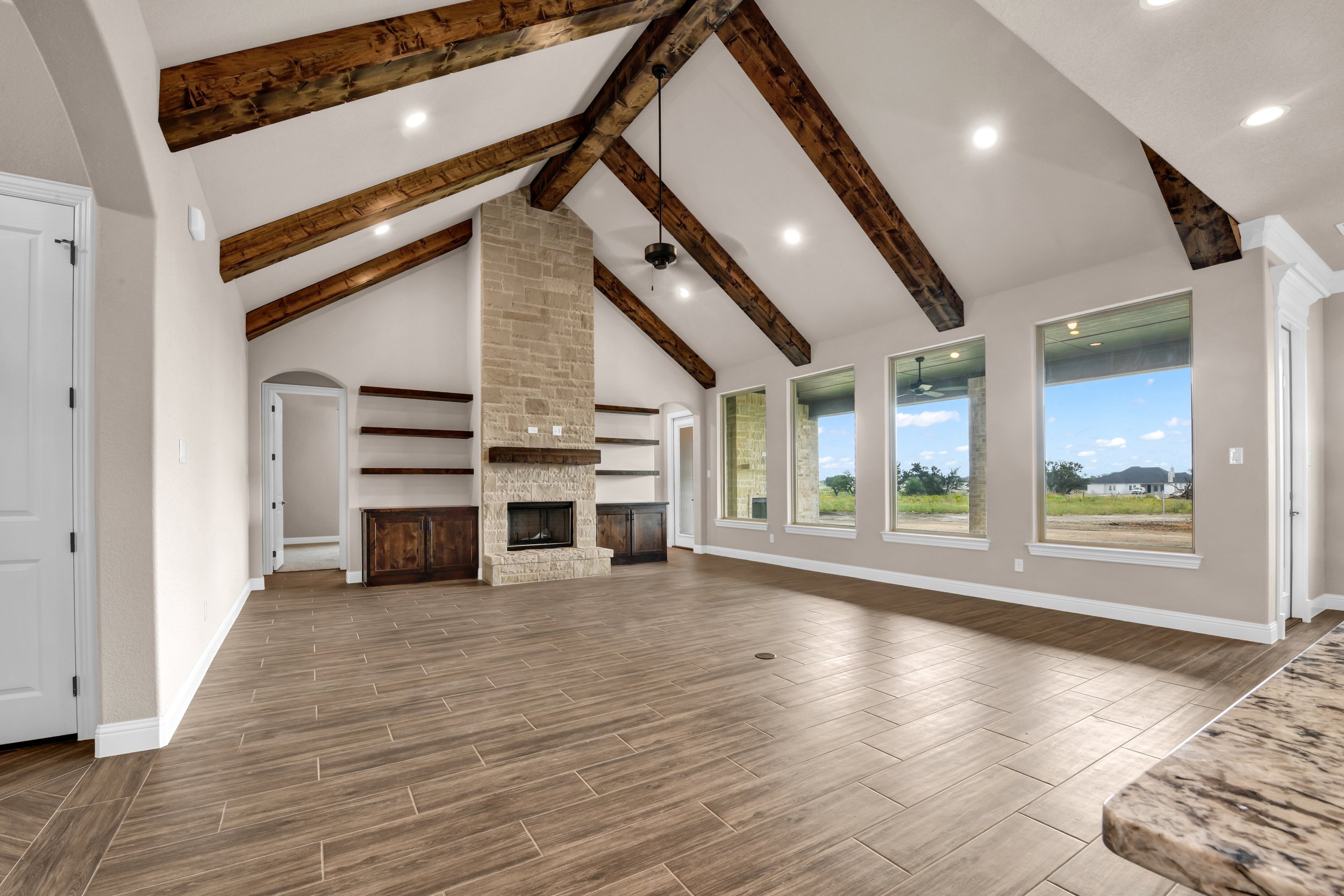 Empty living room with wooden plank flooring, a stone fireplace, built-in shelves, large windows, and wood-beam ceiling.