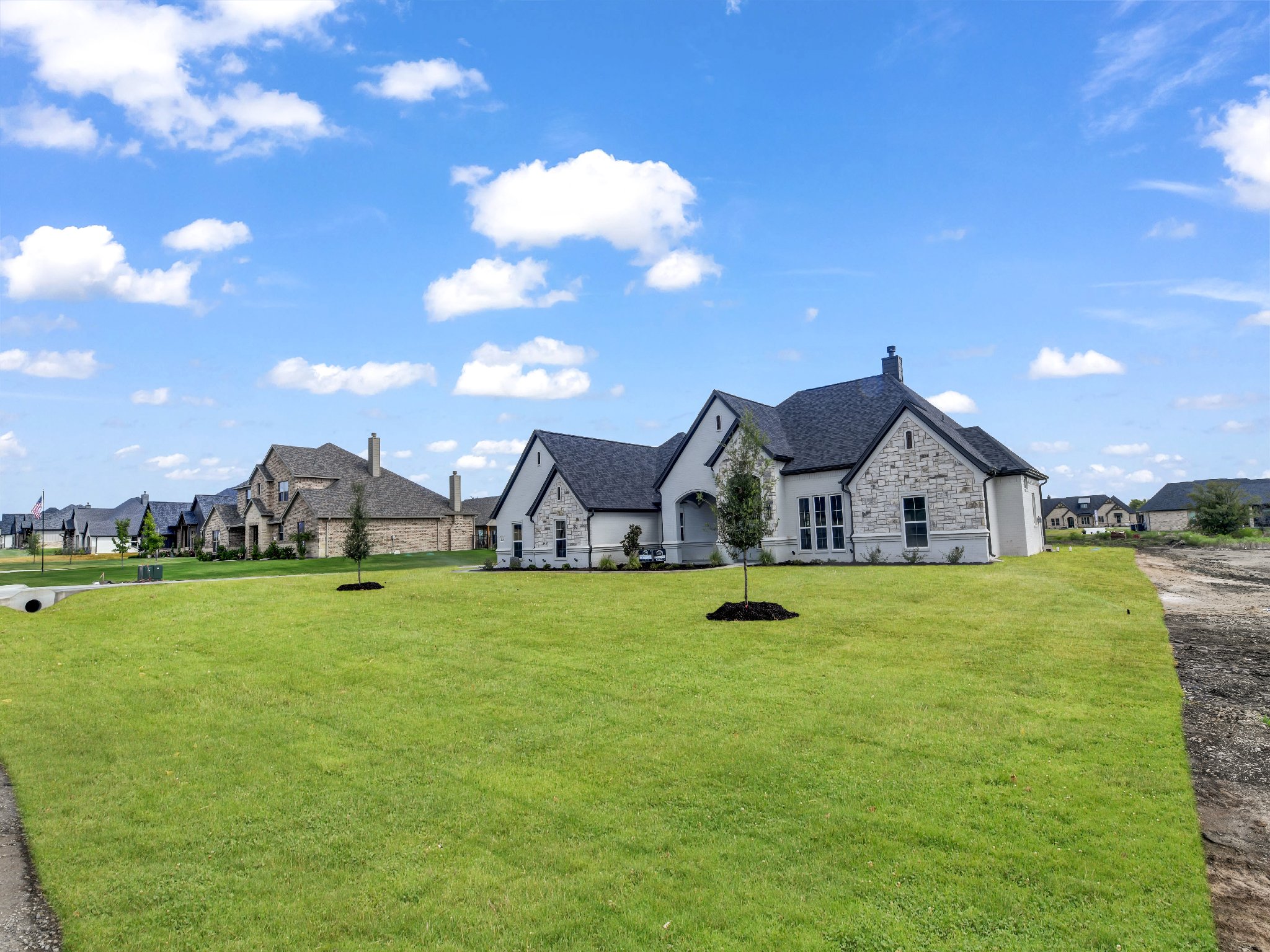 A new white house with stone accents and dark roofing is situated on a green lawn with young trees. Several similar houses are visible in the background under a partly cloudy sky.