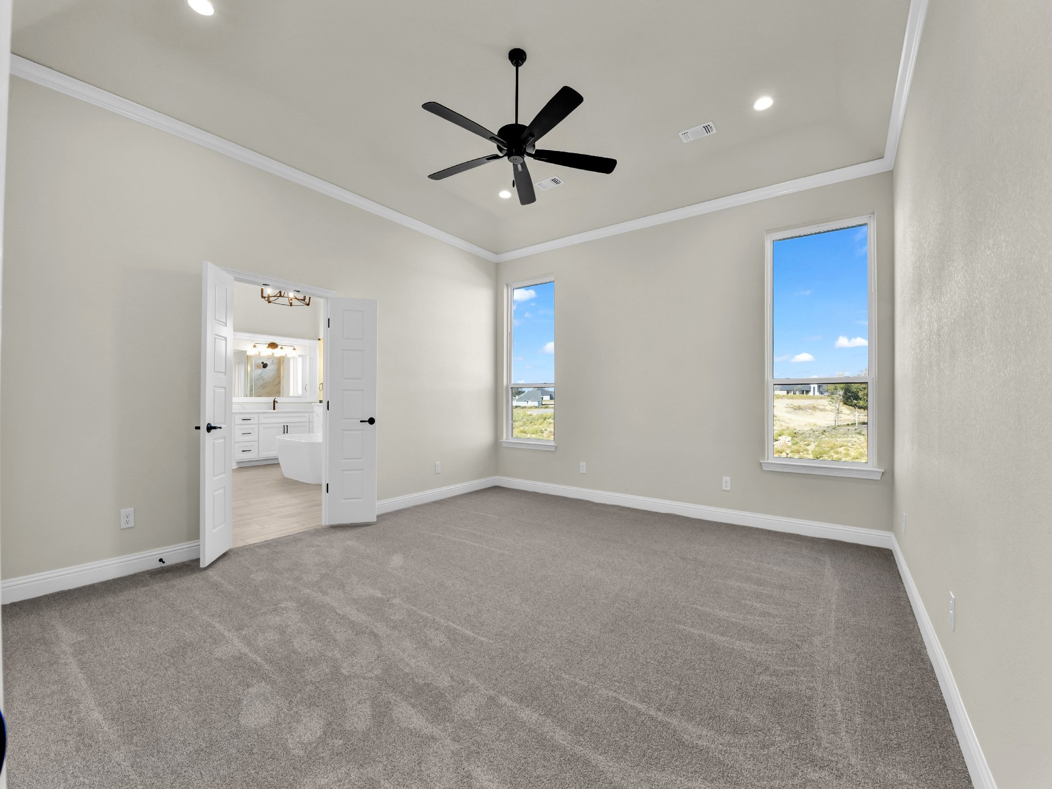 Empty bedroom with beige walls, tan carpet, two windows, white trim, black ceiling fan, and an open doorway leading to a bathroom with white cabinets and bathtub.