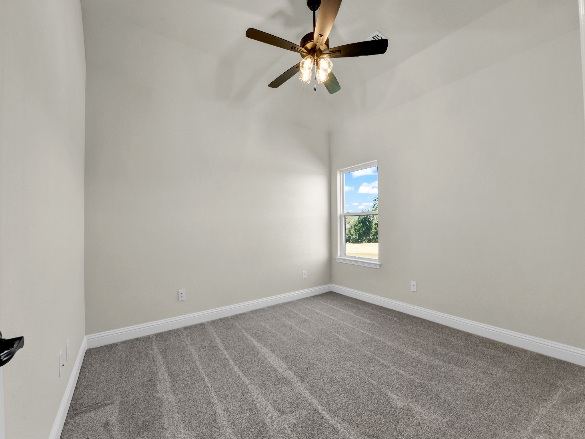 Empty bedroom with beige walls, gray carpet, a window showing a green outdoor landscape, the ceiling fan with lights off, and multiple electrical outlets.