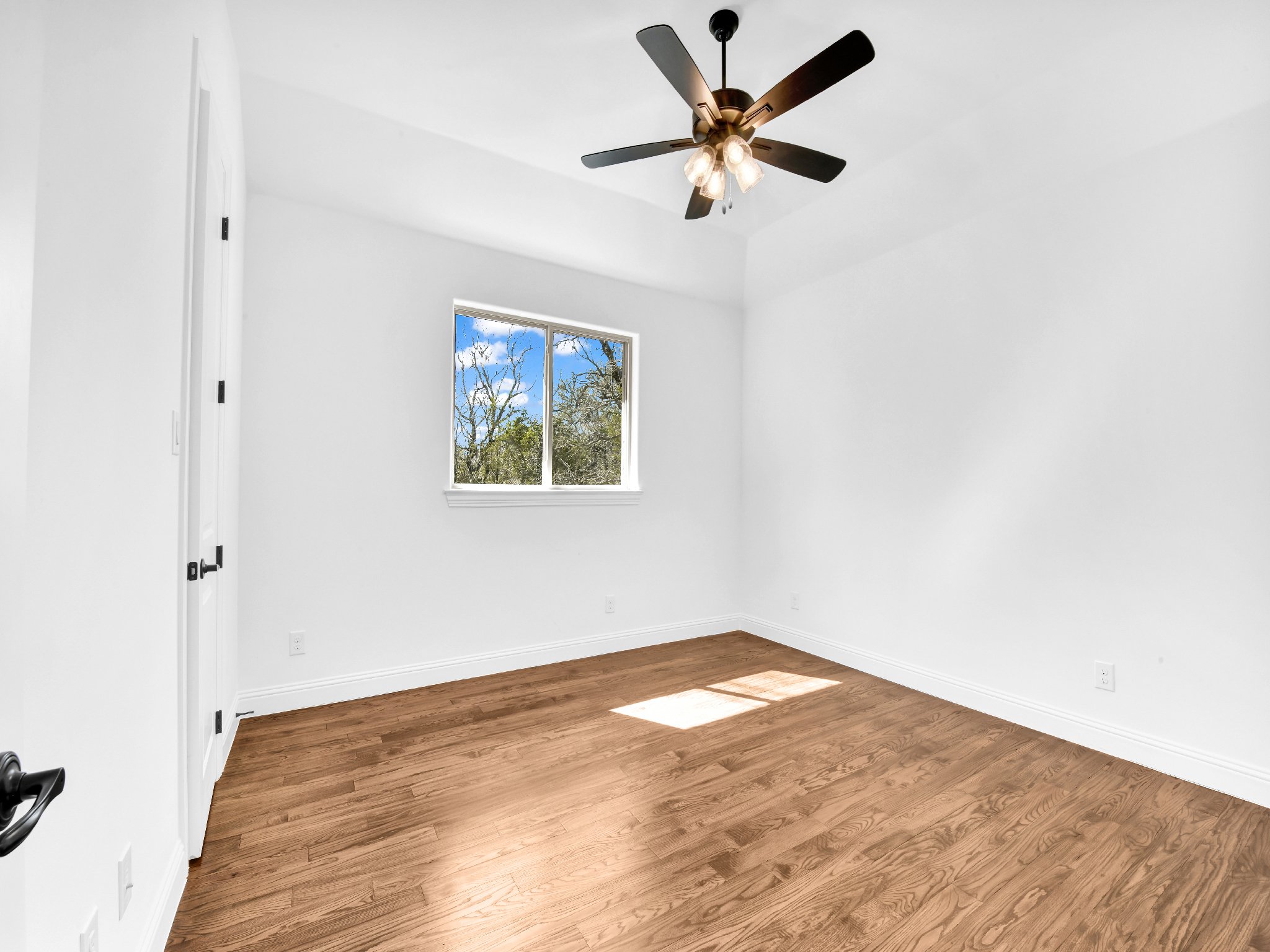 Empty white room with hardwood floors, a window showing trees and blue sky, a ceiling fan with lights, and a closed closet door.