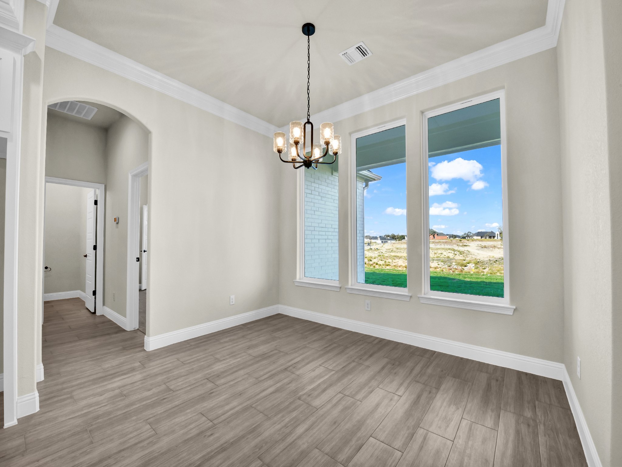 Empty room with three large windows showing a view of a grassy field and houses in the distance, chandelier hanging from the ceiling, white walls, crown molding, and light wood flooring.