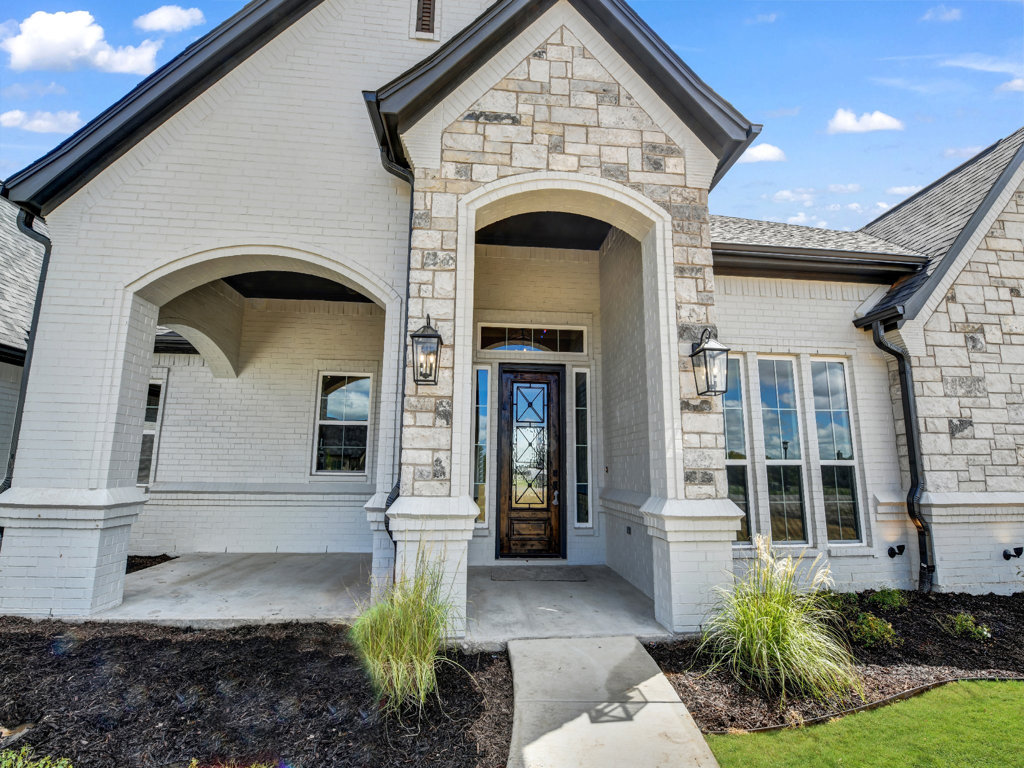 Front entrance of a modern house with white brick and stone exterior, black front door with glass panels, two outdoor lanterns, and landscaped garden with grass and plants.