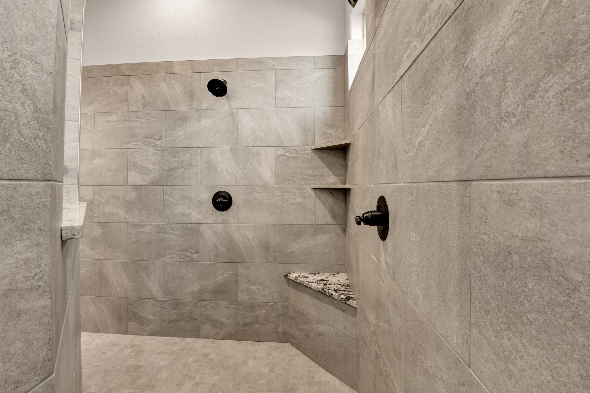 A modern walk-in shower with gray tiled walls, a marble shelf, and black fixtures, including a showerhead and controls.