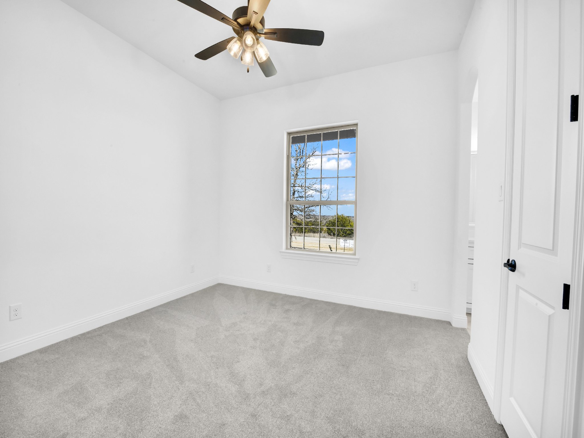 Empty bedroom with white walls, a window showing a blue sky with clouds, beige carpet, a ceiling fan with lights, and a white door.