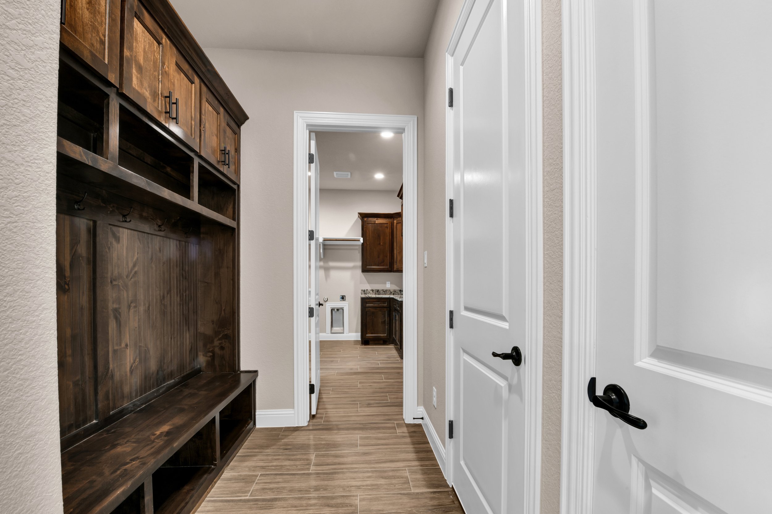 A hallway with light-colored walls, brown wood flooring, a white door on the right, a dark wood coat rack on the left, and a laundry area visible at the end of the hall.