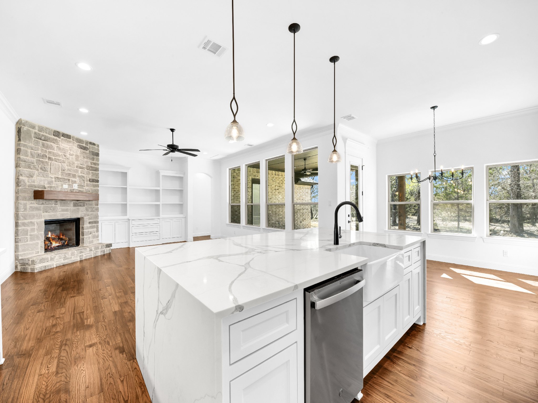 Bright, open-concept living area with white walls, a stone fireplace, wooden flooring, large windows, and a kitchen island with a marble countertop, black faucet, and pendant lights.