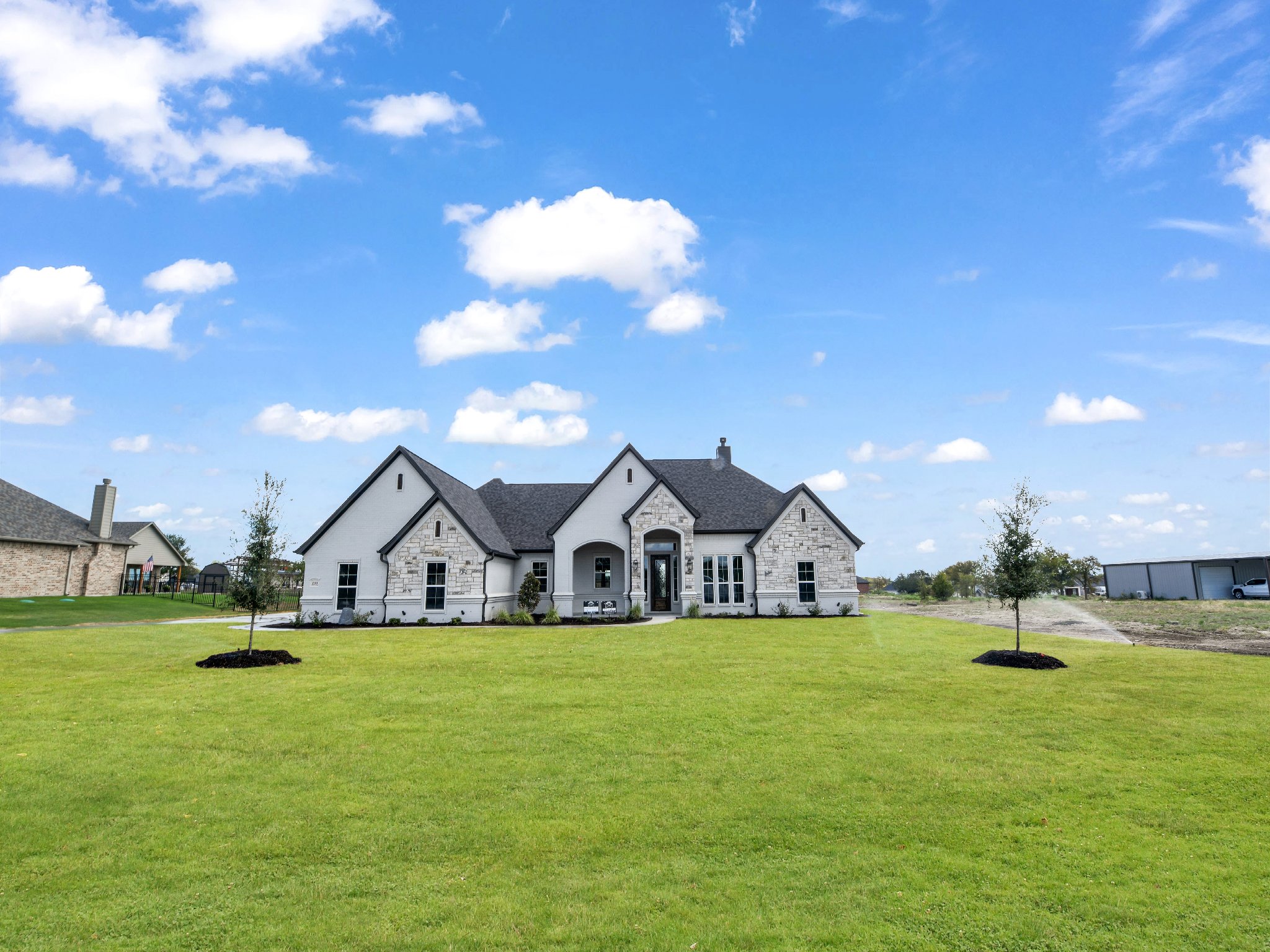 A modern white house with stone accents and a dark roof, surrounded by a green lawn and two small trees under a partly cloudy blue sky.