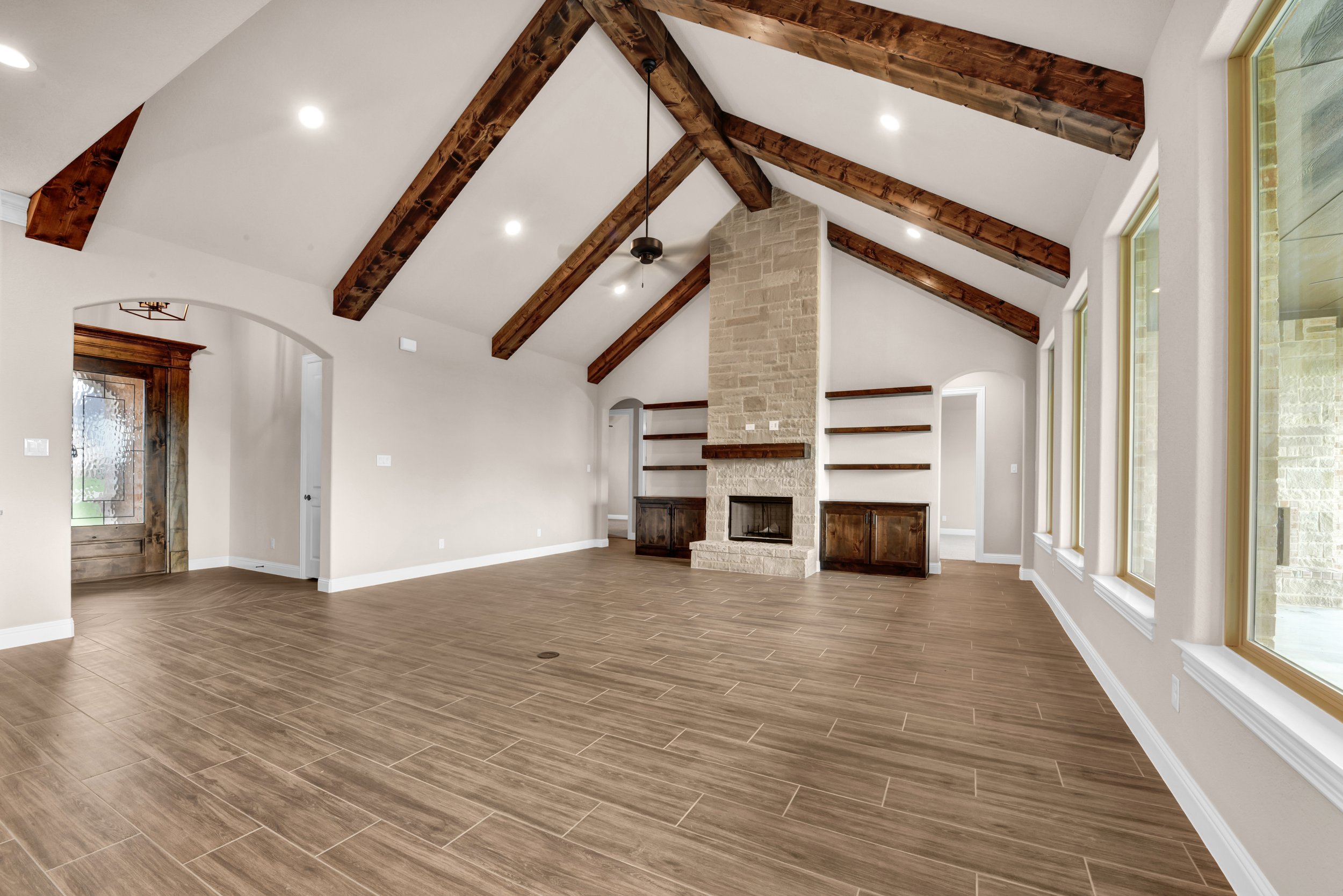Empty living room with vaulted ceiling, exposed wooden beams, a stone fireplace, large windows, and built-in wooden shelves and cabinets.