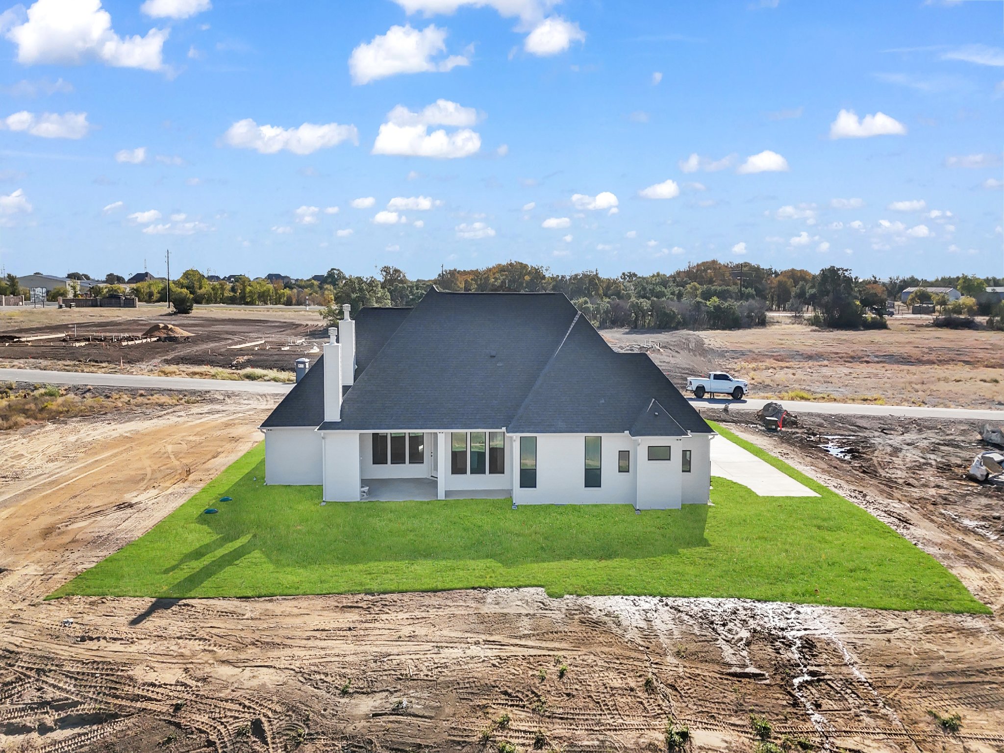 A newly built white house with a dark roof on a grassy lot, under a partly cloudy sky, with a dirt area around it and construction vehicles in the background.