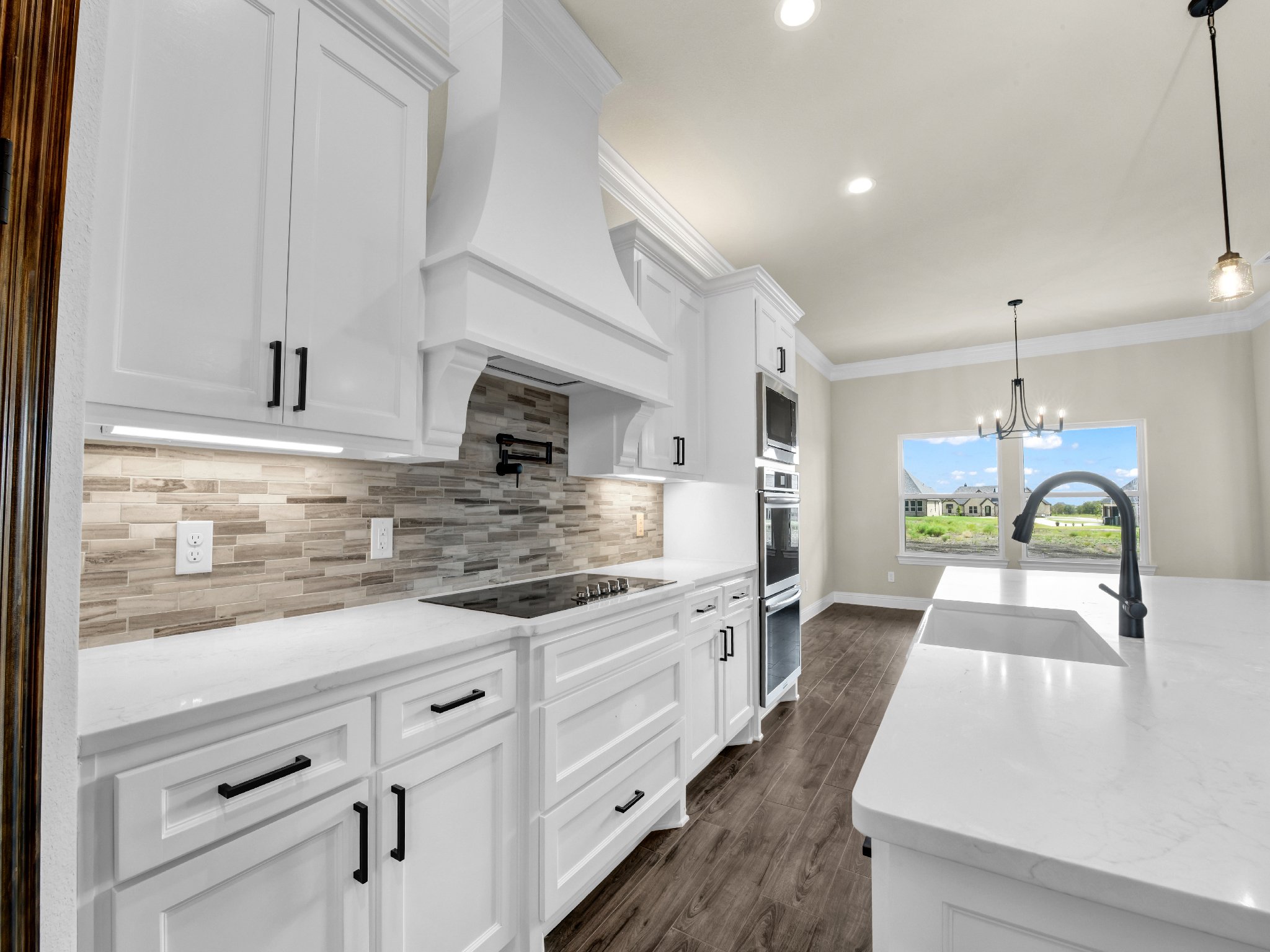 Modern kitchen with white cabinets, a marble countertop, a mosaic tile backsplash, and a double oven, overlooking a dining area with a chandelier and large window showing a sunny outdoor scene.