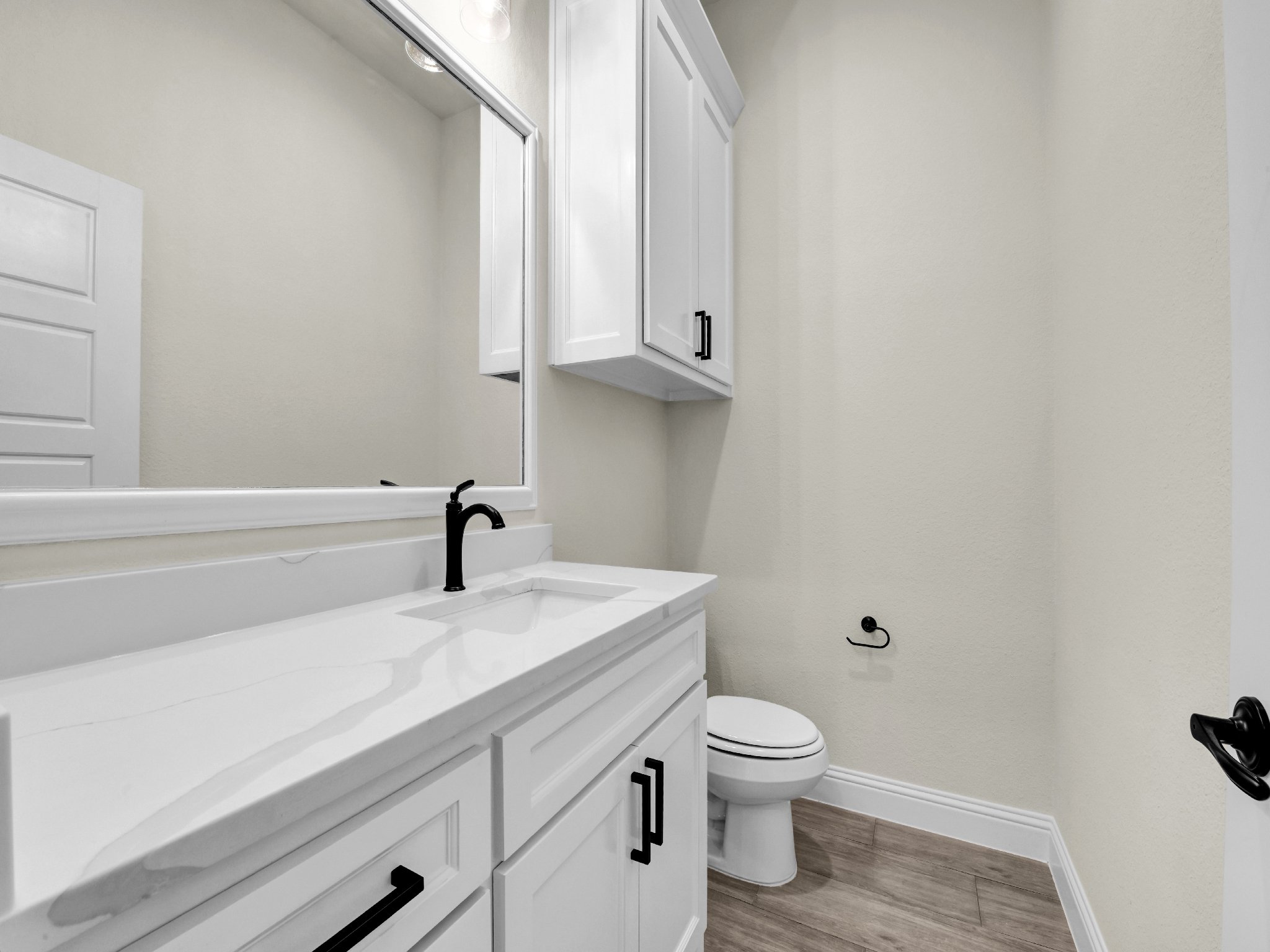 A small, modern bathroom with white cabinetry, a white countertop with a black faucet, a large mirror, a wall-mounted cabinet, a toilet, and wood-like flooring.