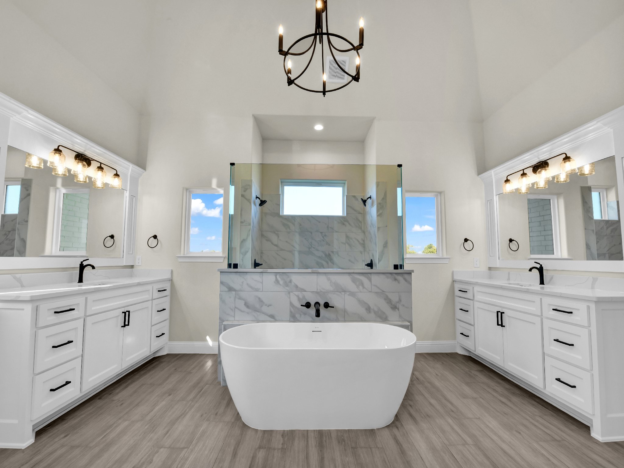 Modern, spacious bathroom with dual vanities, large mirrors, black fixtures, a freestanding tub, and a glass-enclosed shower with marble tiles, topped by a black chandelier, and windows showing a blue sky with clouds.