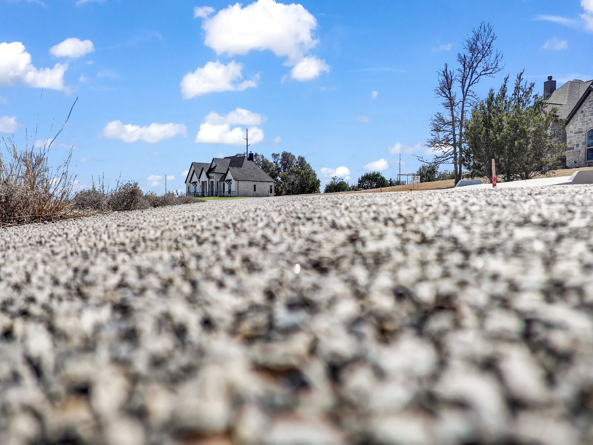 Low angle view of a textured asphalt road with houses, trees, and a blue sky with scattered clouds in the background.