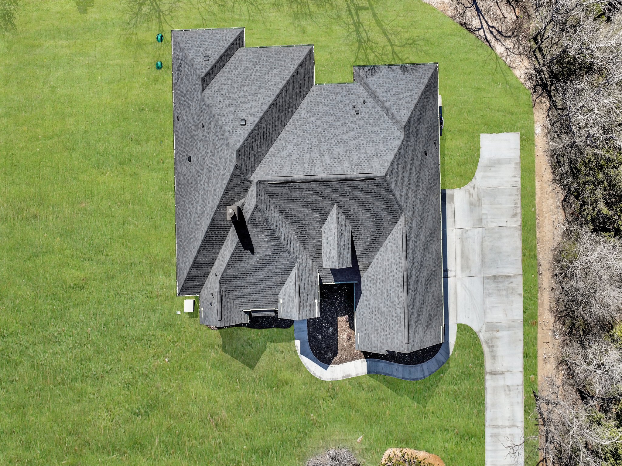 Aerial view of a house with a gray shingled roof, surrounded by green yard and trees, with a concrete driveway leading to the house and a curved walkway.