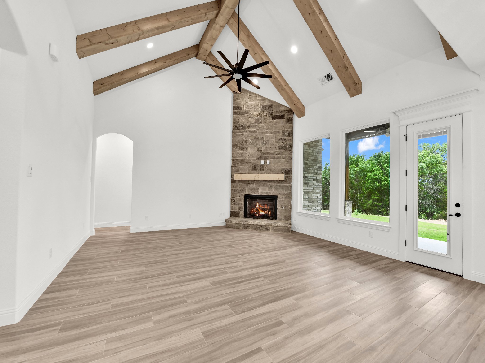 Empty living room with white walls, wooden beams on the ceiling, a stone fireplace, large windows, and a glass door leading outside
