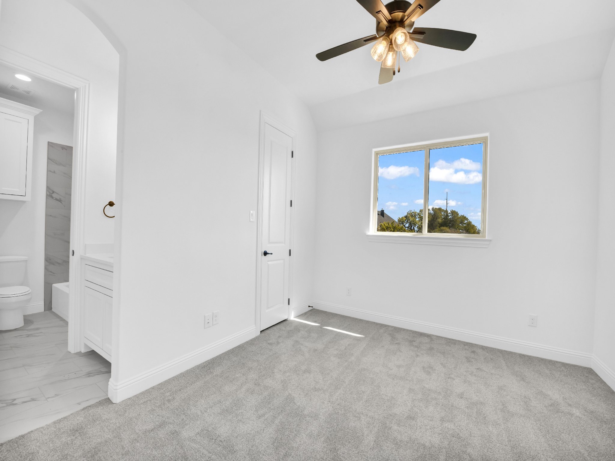 Empty white bedroom with gray carpet, ceiling fan with light, window showing blue sky and trees, doorway to bathroom.