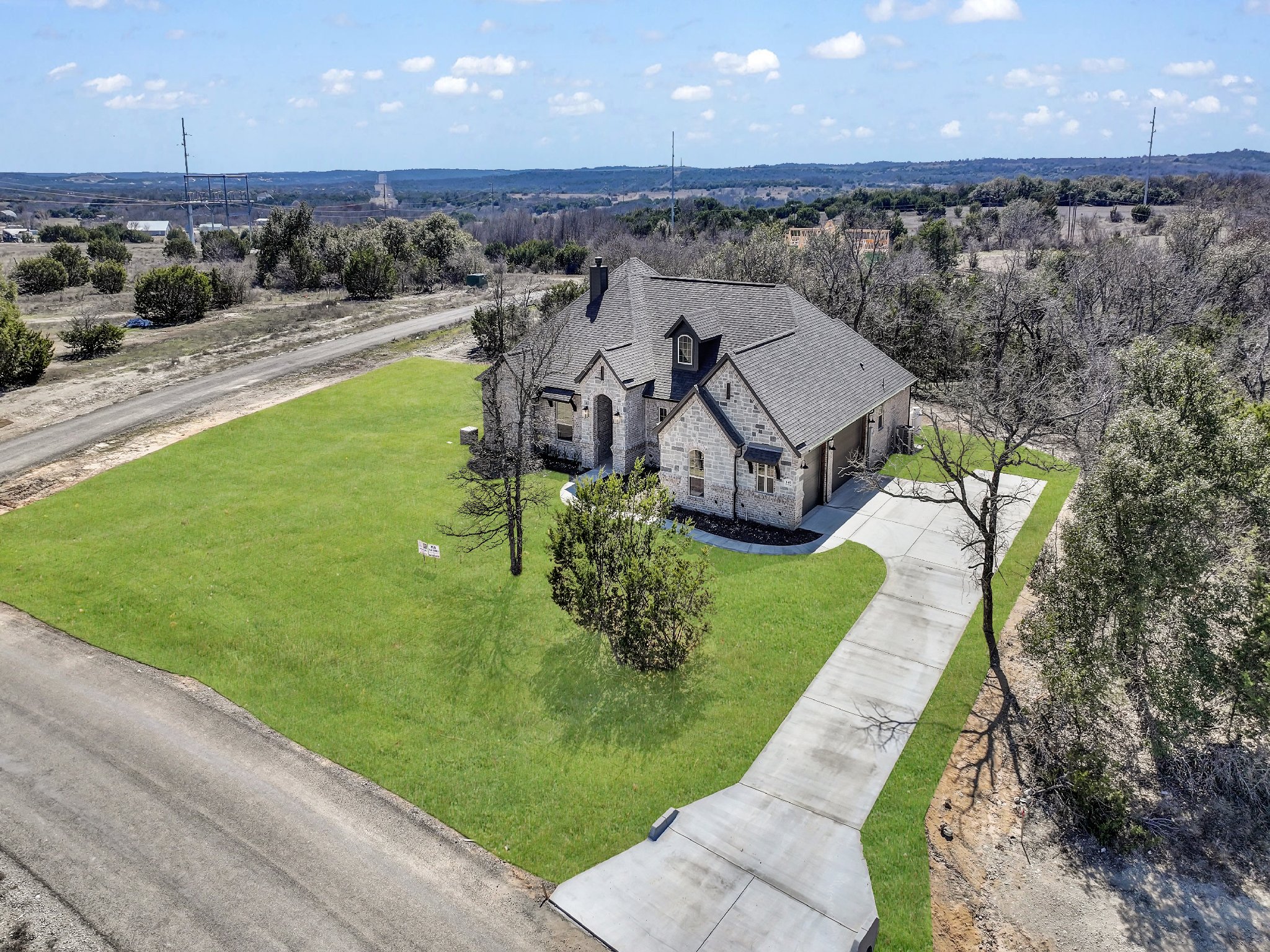A large house with brick and stone exterior, surrounded by a green lawn and leafless trees. The house has a curved driveway leading to a two-car garage. The surrounding area appears semi-rural with a dirt road nearby and power lines in the distance.