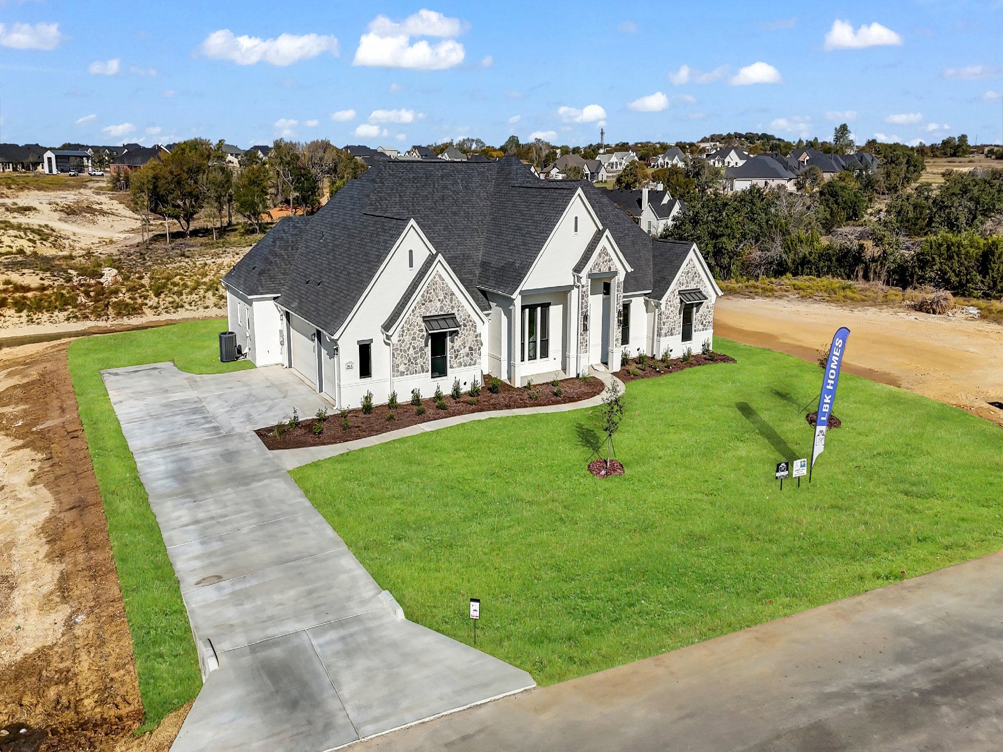 New white house with black roof and stone accents, green lawn, and driveway, in a suburban area with other houses in the background.