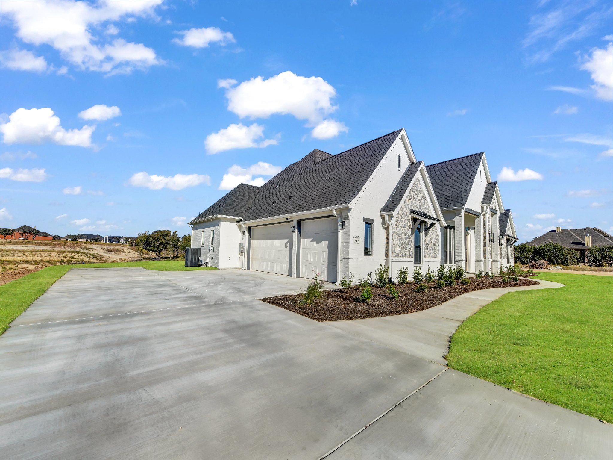 Modern white house with a stone accent, multiple gable roofs, and a driveway leading to a two-car garage, set against a bright blue sky with scattered clouds.