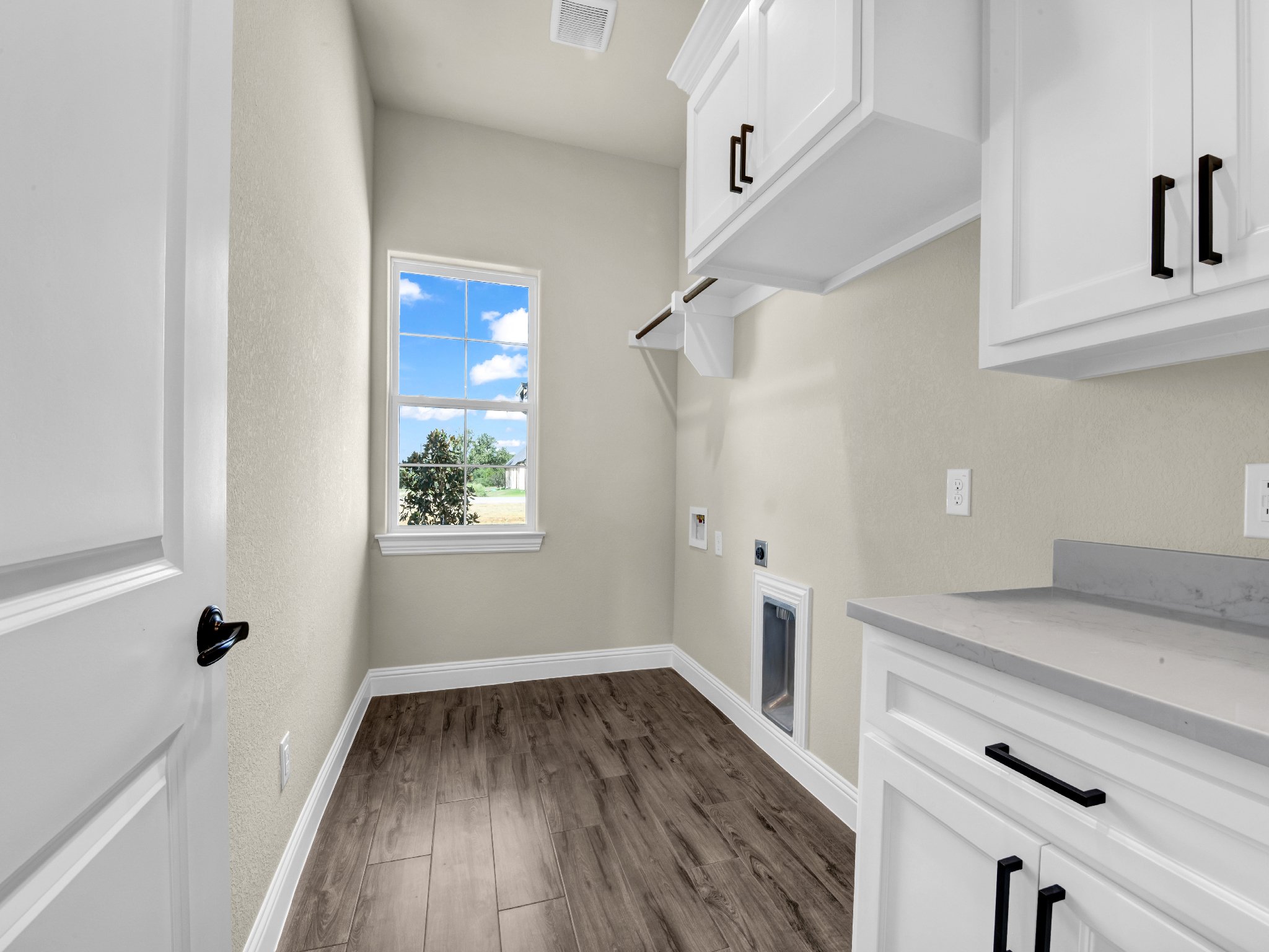 Empty laundry room with white cabinets, a window with a view of green trees outside, and dark wood flooring.
