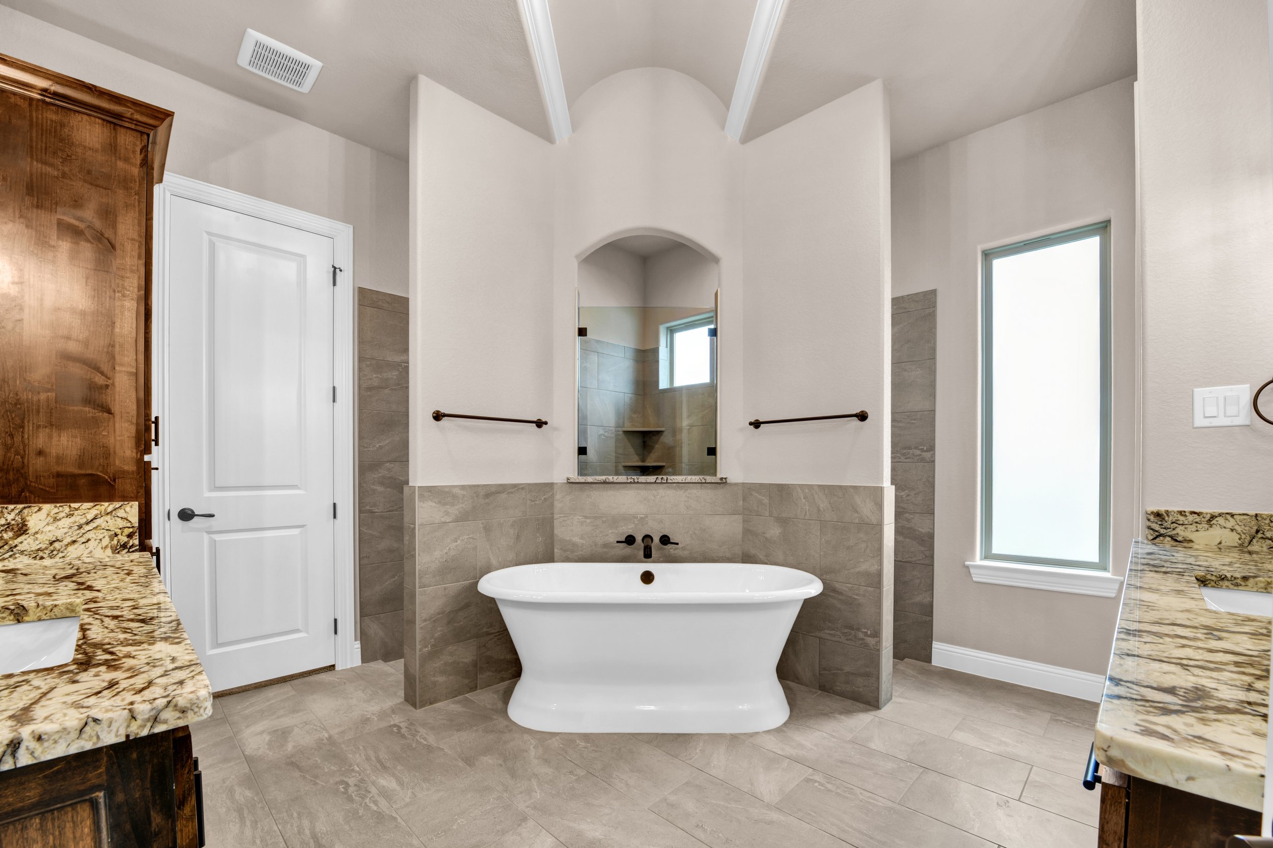 Modern bathroom with a white freestanding bathtub, beige tiles on the lower walls and floor, granite countertops, and two windows with frosted glass.