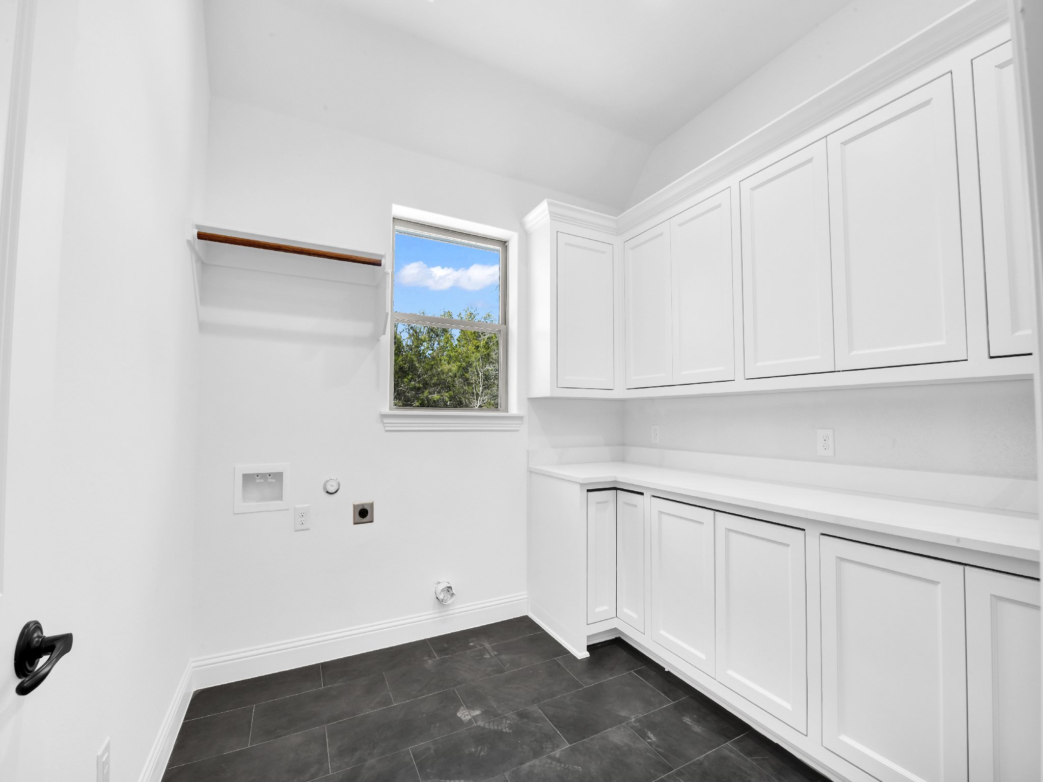 Empty laundry room with white cabinets, a window showing blue sky and trees, and a dark tiled floor.