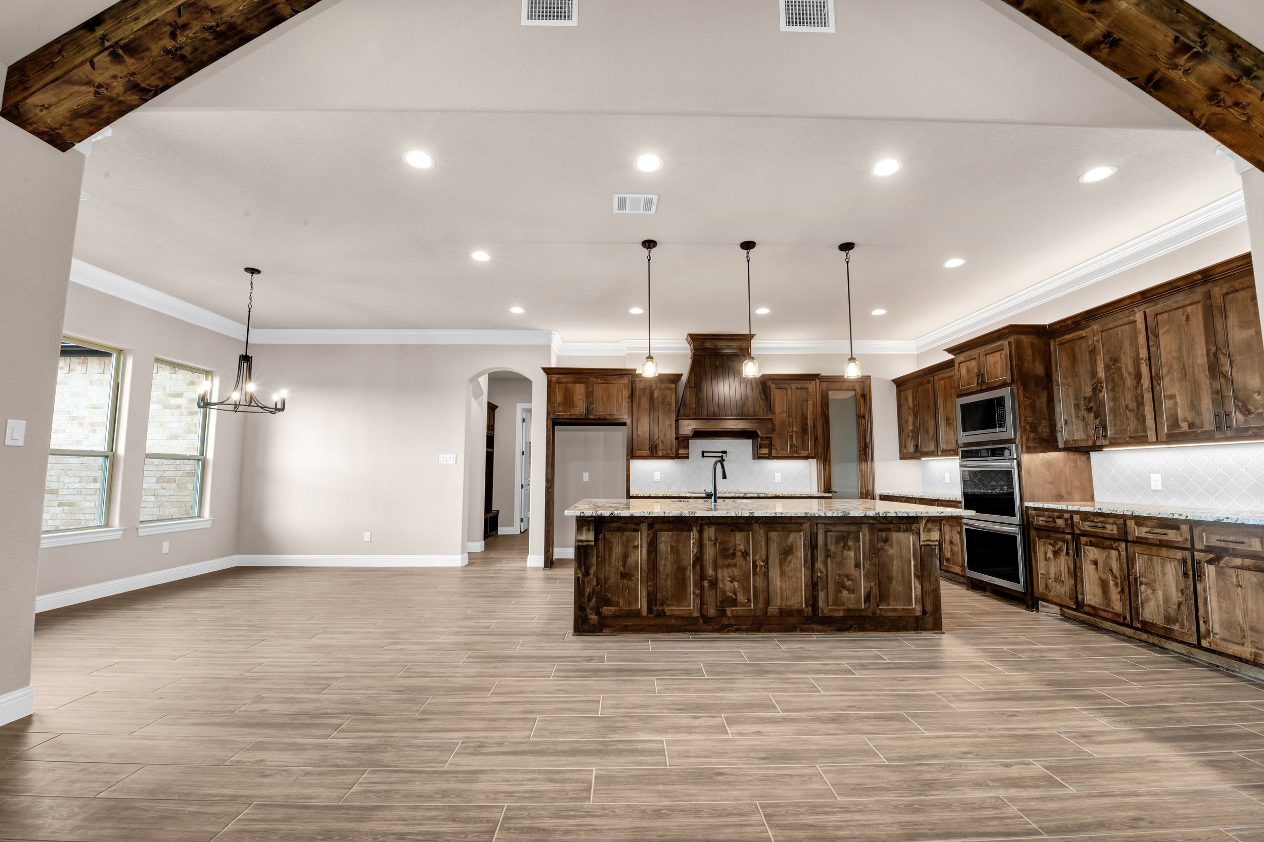 Spacious kitchen with wooden cabinets, a kitchen island with granite countertop, modern appliances, and pendant lighting, open to a dining area with a chandelier, hardwood flooring, and windows with brick walls outside.