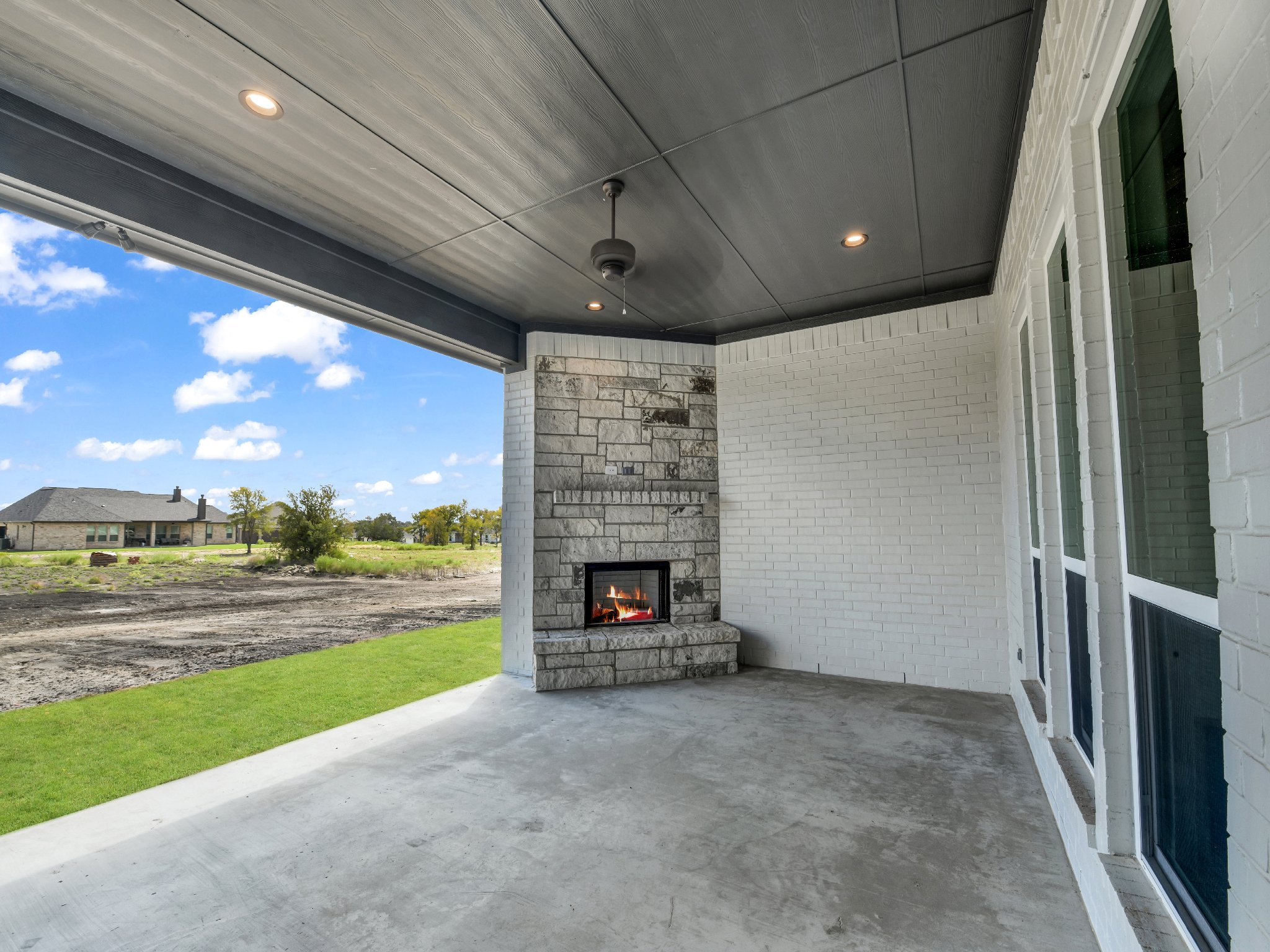 Covered porch area with a stone fireplace, ceiling fan, ceiling lights, white brick walls, and a view of a grassy yard and neighboring houses in the distance.