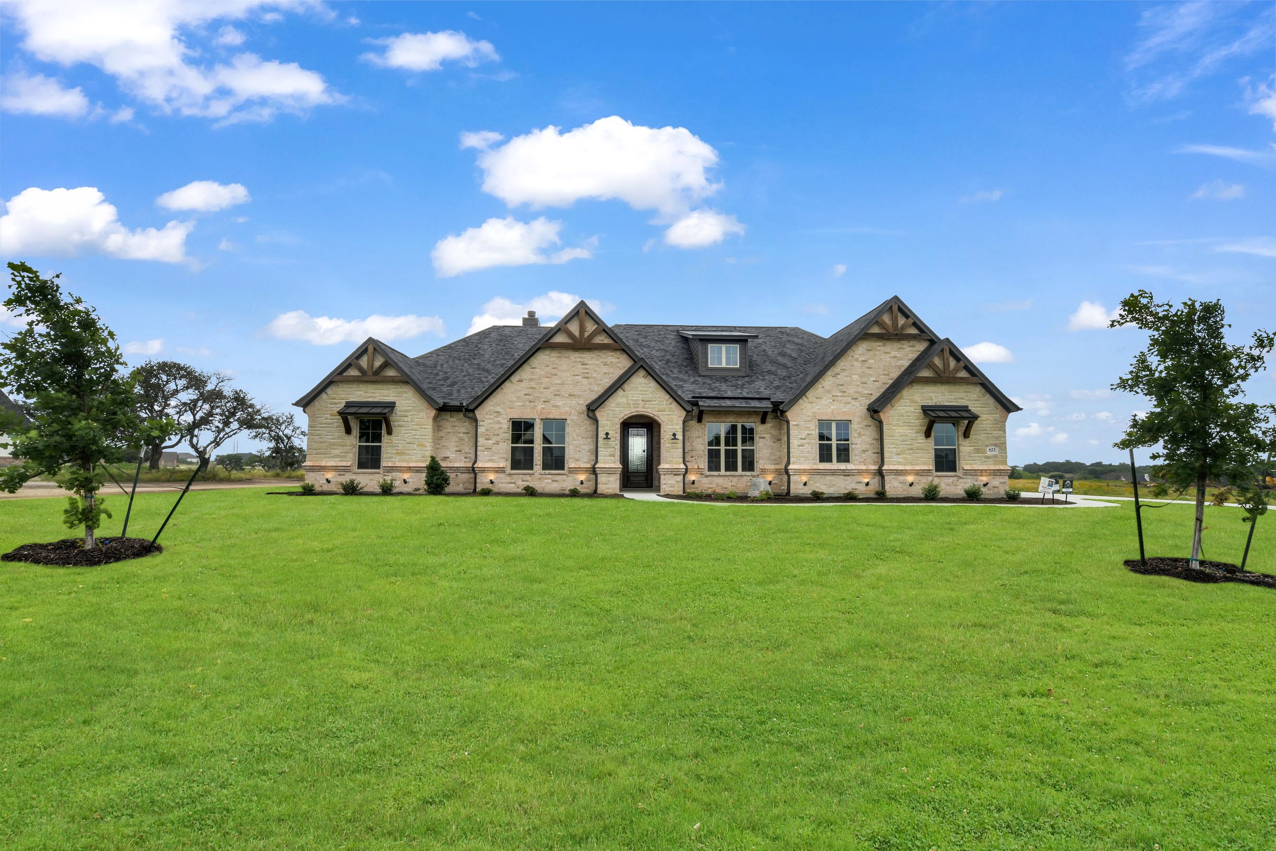 A large, modern house with stone exterior walls, dark roof, surrounded by green lawn and trees under a blue sky with clouds.