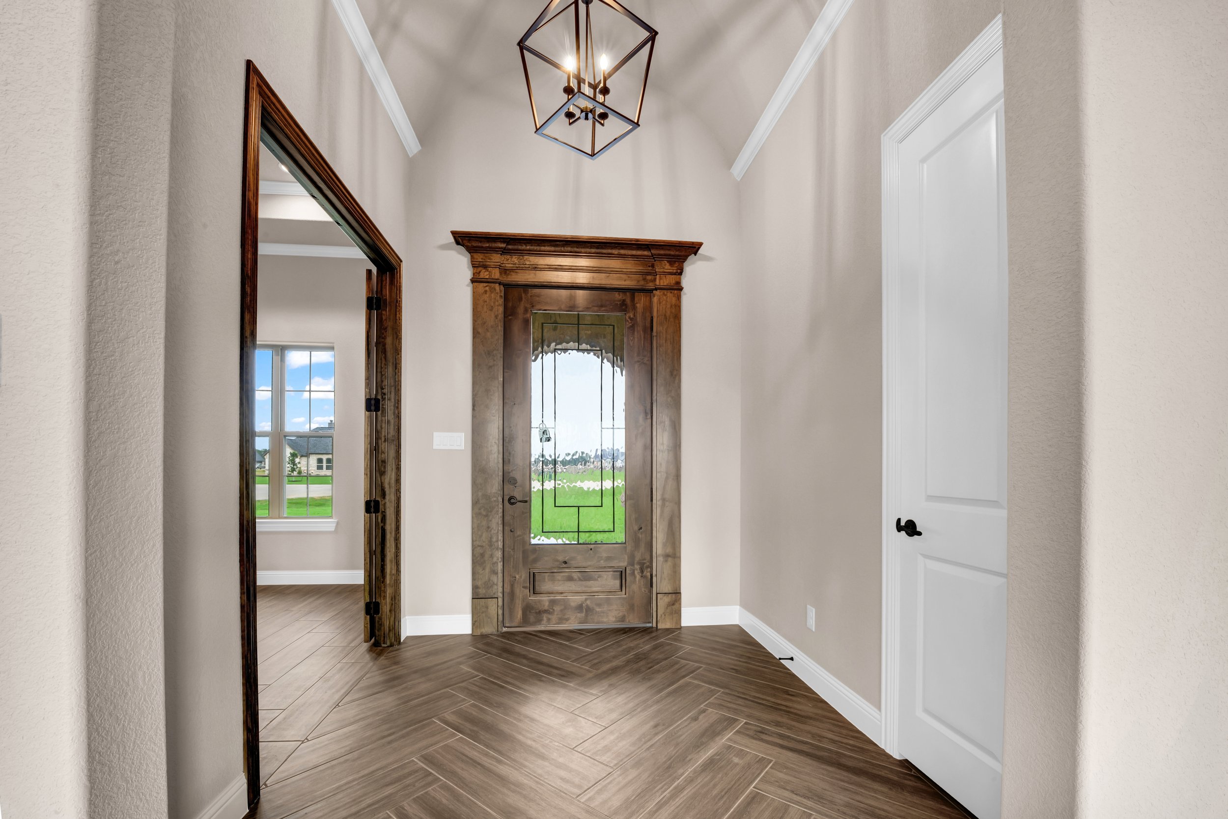 Entryway with a wooden front door with decorative glass panel, beige walls, white crown molding, a modern geometric chandelier, and a view of a green lawn and neighboring houses through the open door and windows.