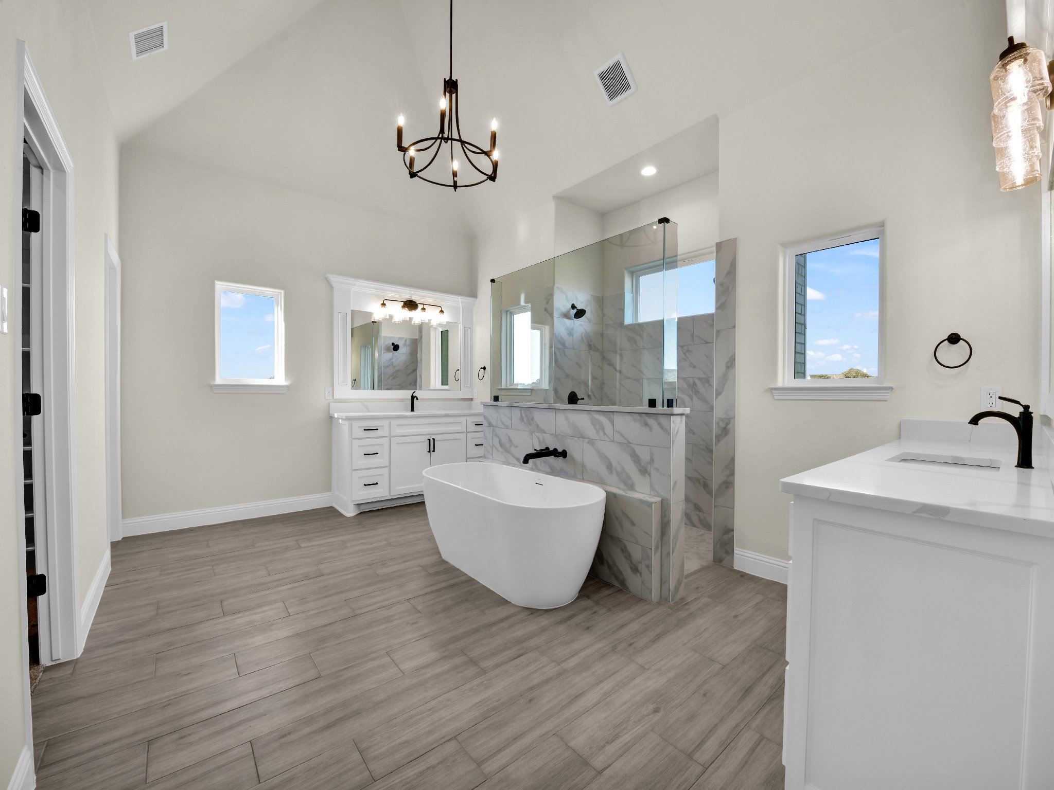 Modern bathroom with freestanding bathtub, marble shower, white cabinetry, wood-look flooring, and windows showing a blue sky.